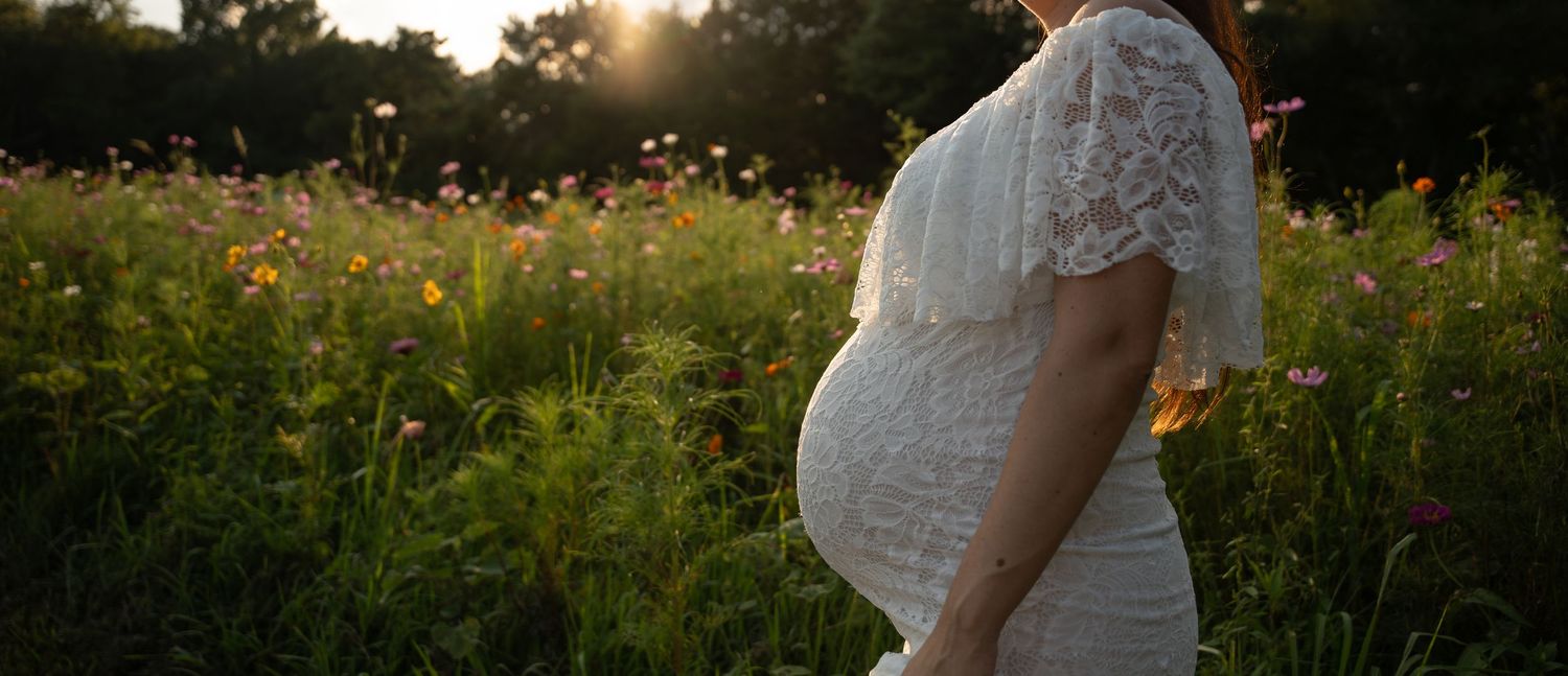 Pregnant woman in white lace dress standing in colorful wildflower meadow at sunset.