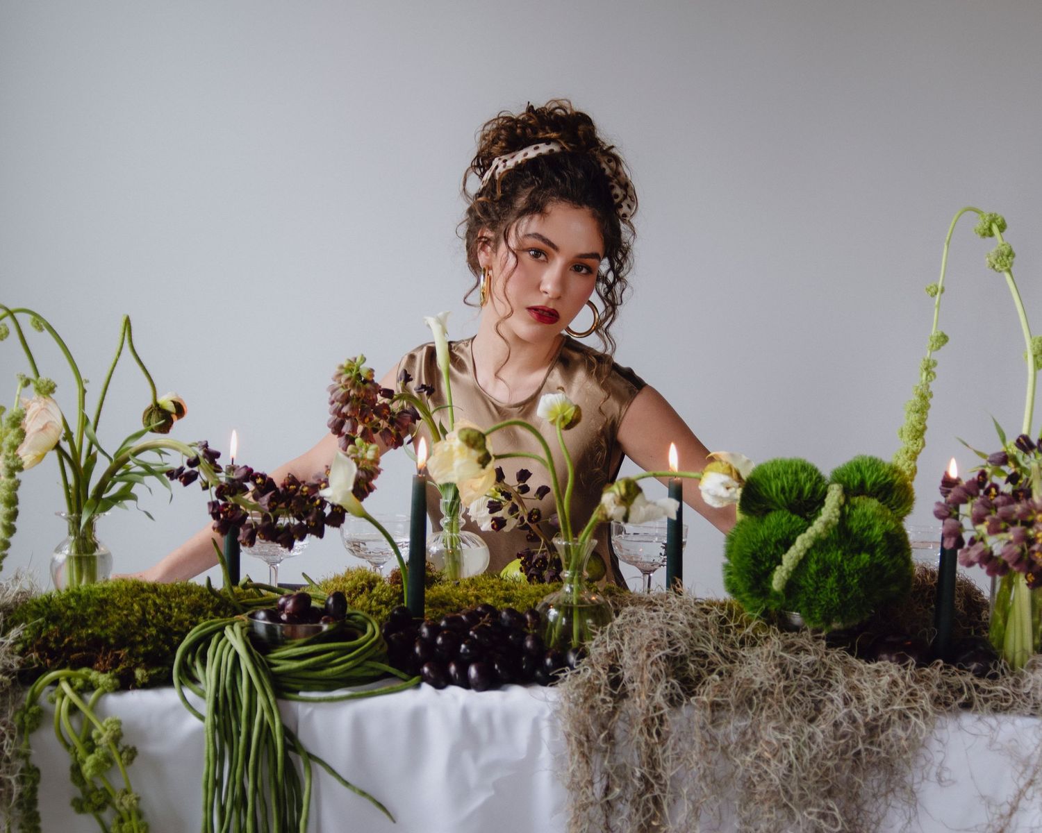 Young woman with updo arranges exotic floral centerpiece with moss and dark flowers on white table.