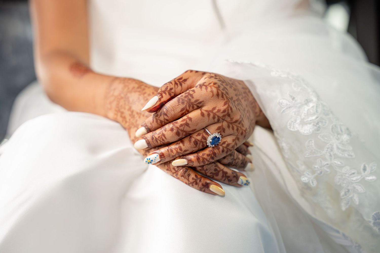 Close-up of bride's henna-adorned hands with blue sapphire ring and decorated nails on white wedding dress.