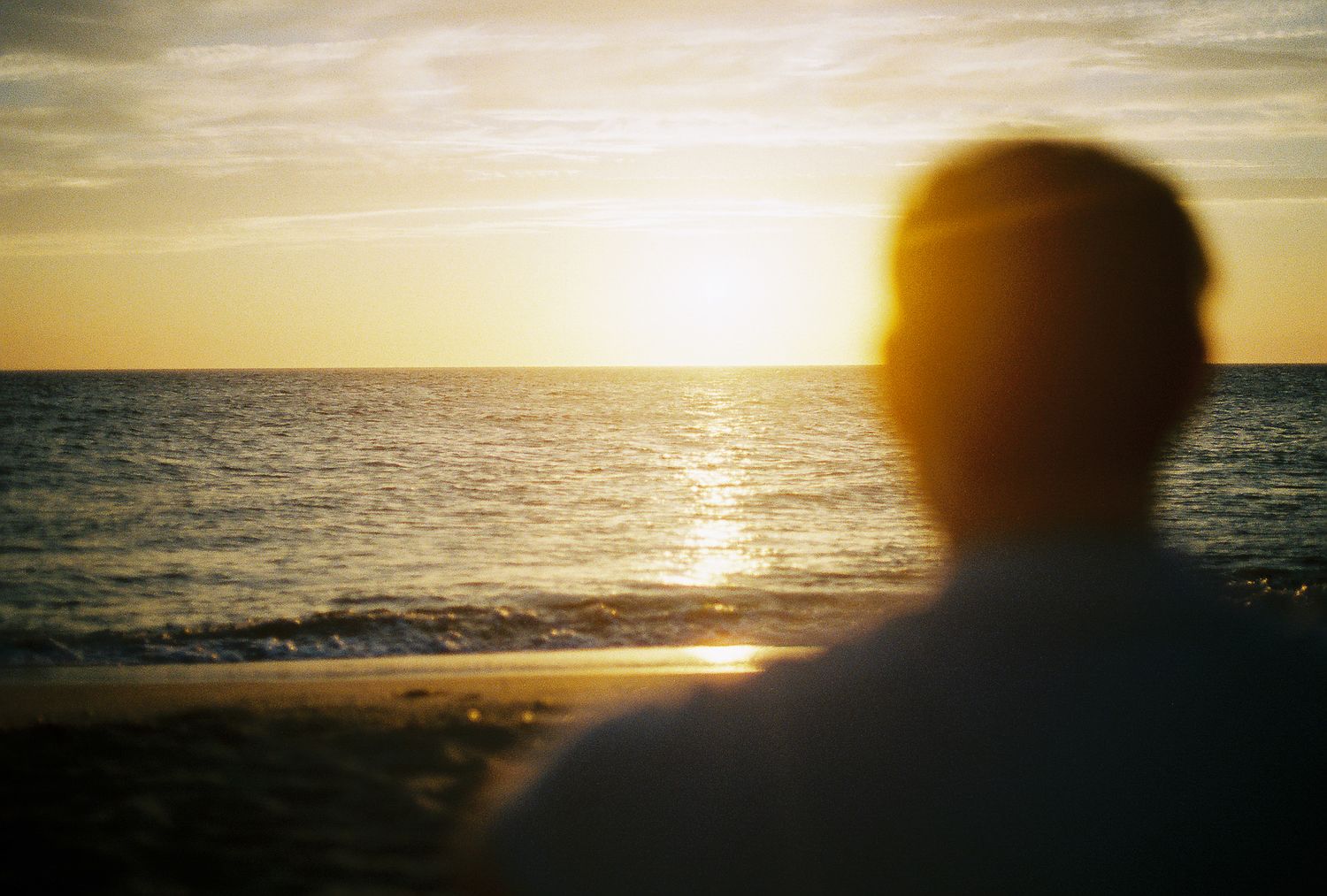 Silhouette of person watching golden sunset over calm ocean waters from sandy beach.