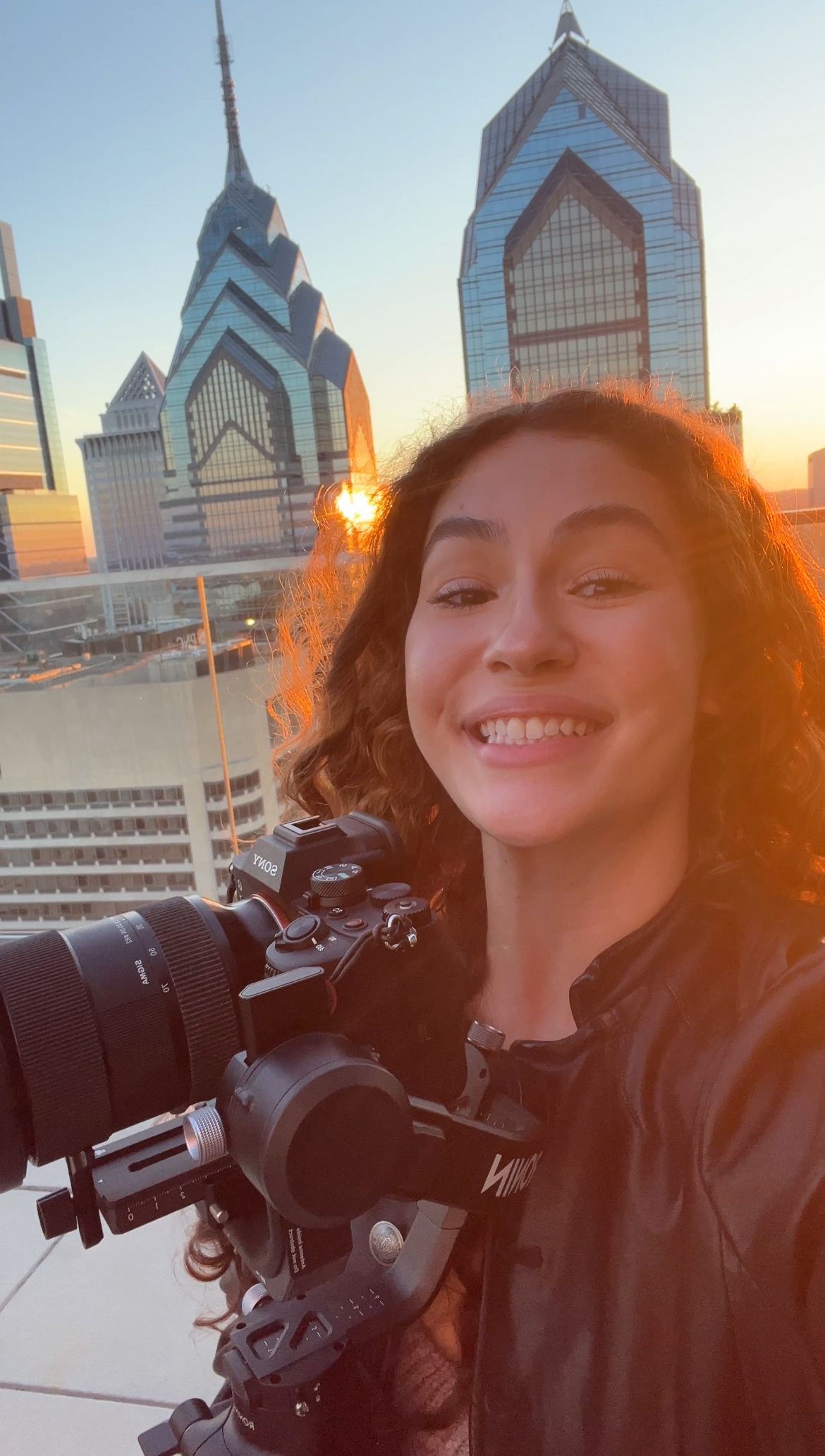 Young woman smiling with camera on gimbal against Philadelphia skyline at sunset.