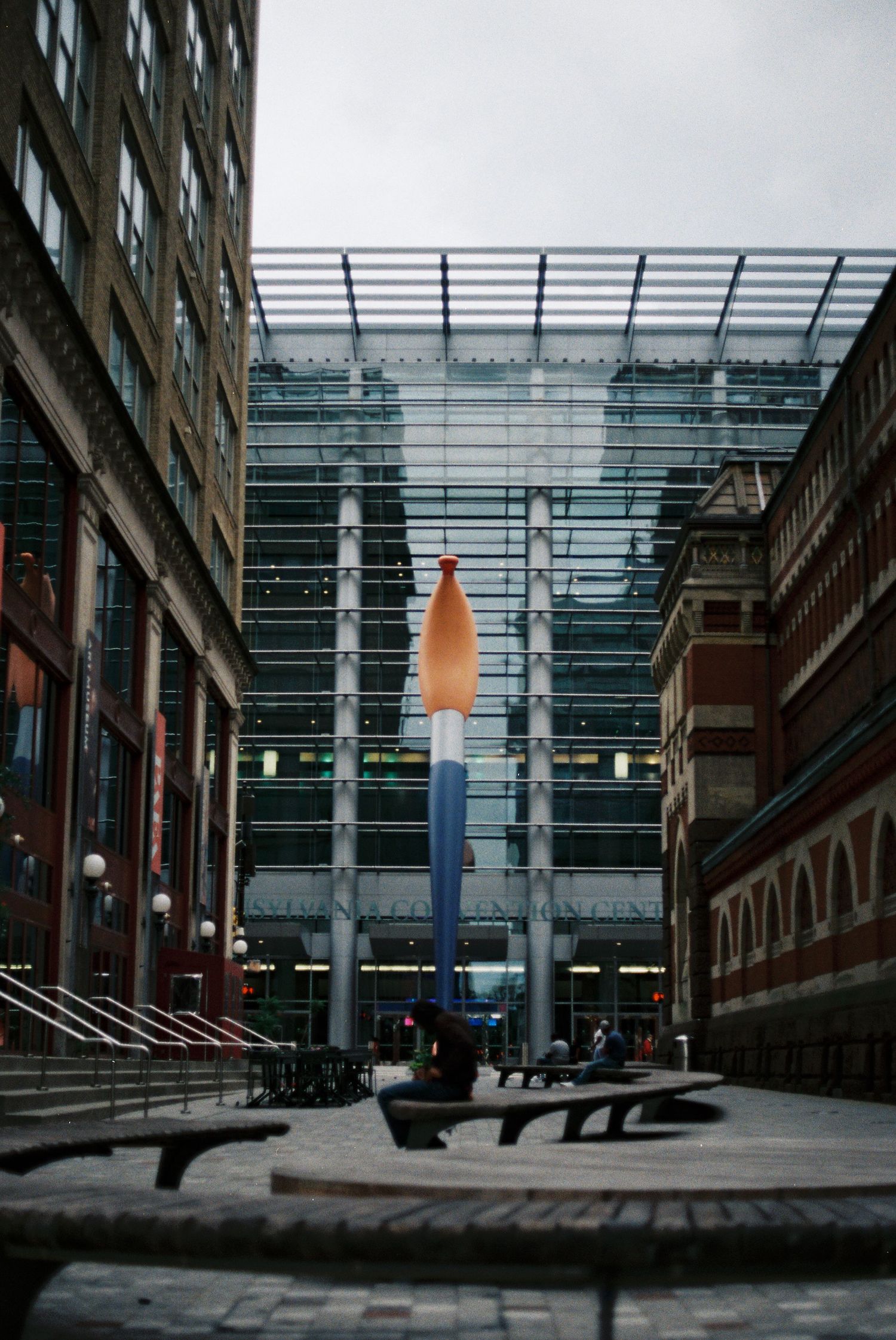 Colorful rocket sculpture displayed in a modern glass atrium courtyard between brick buildings.