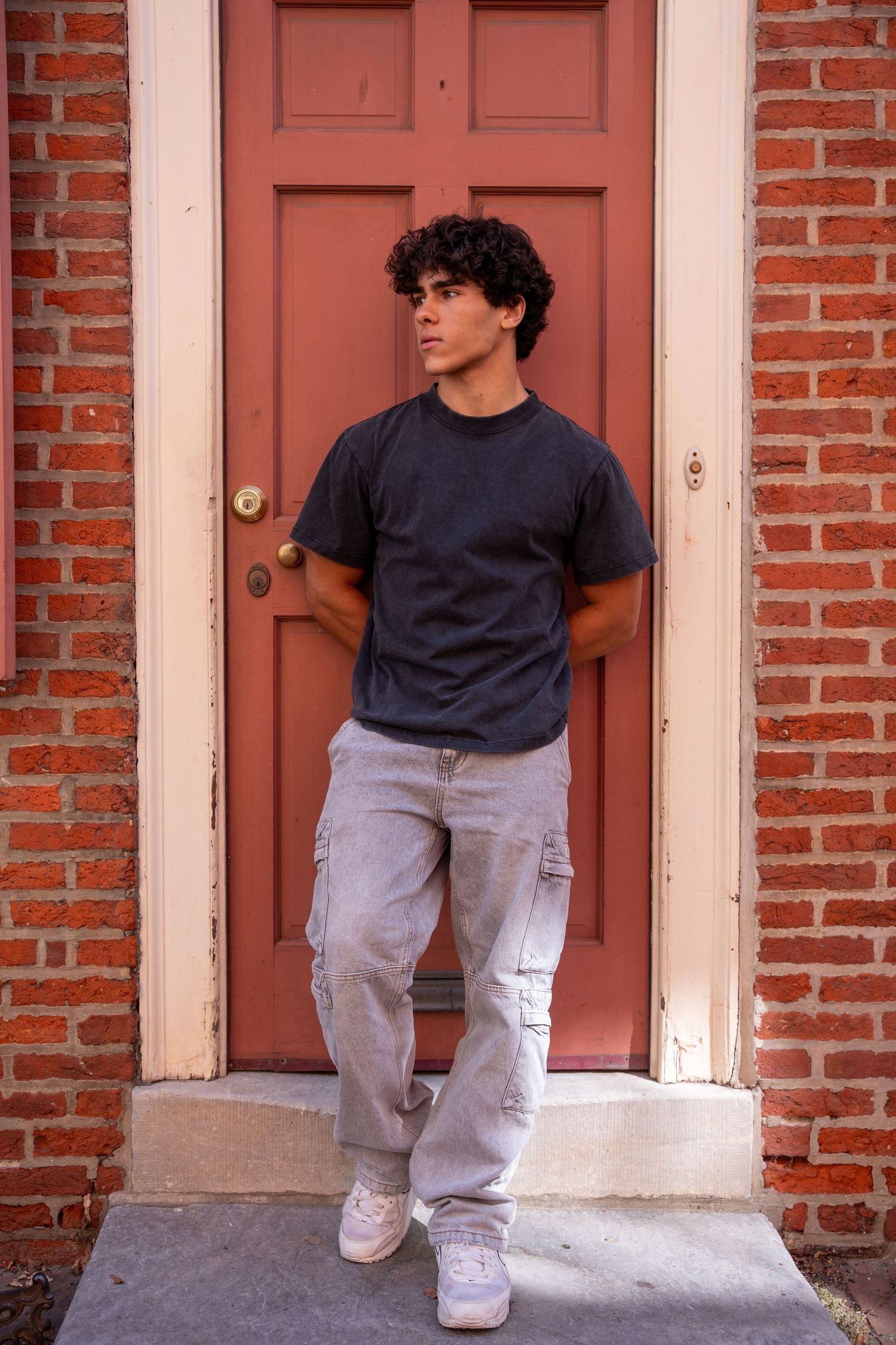Young man with curly hair in black tee and cargo pants standing by red brick doorway.