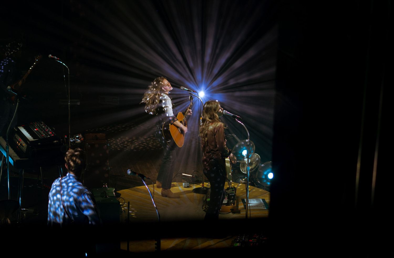 Female guitarist performing on a dimly lit stage with a bright starburst light behind her head.