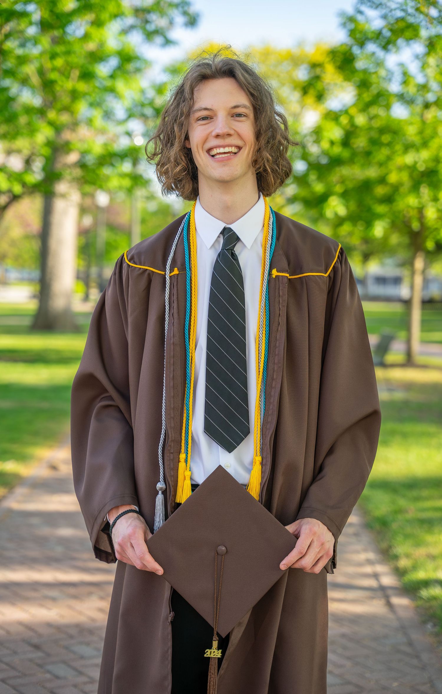 Smiling male graduate in brown cap and gown with colorful stoles on campus.