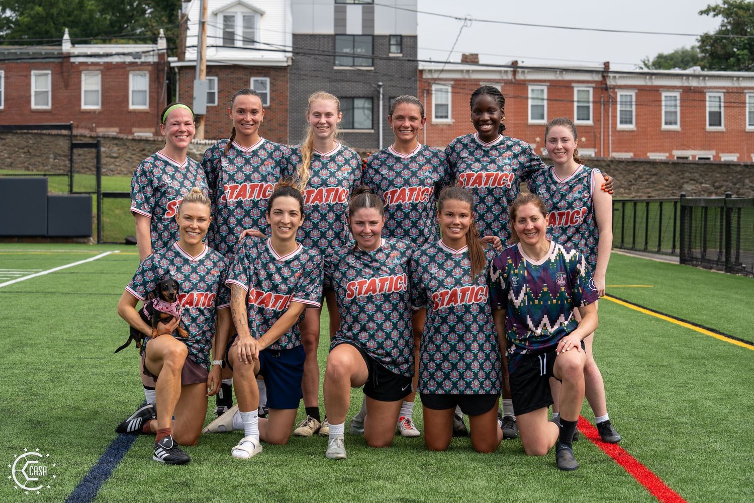 Women's flag football team poses together on a green turf field wearing matching gray jerseys outdoors.