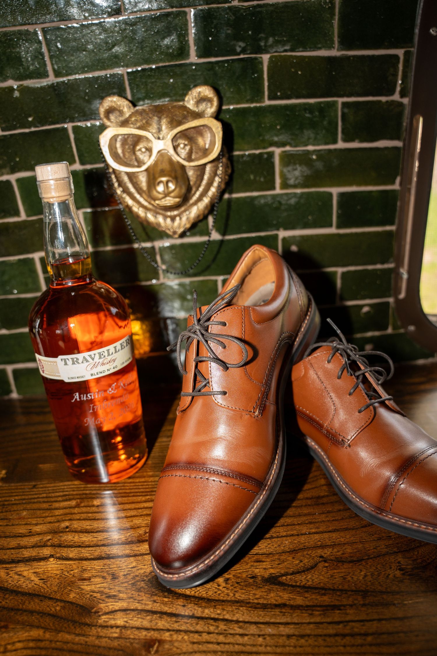 Brown leather cap-toe dress shoes beside a bottle of Traveller whiskey, with a gold bear wall mount on green tiles.