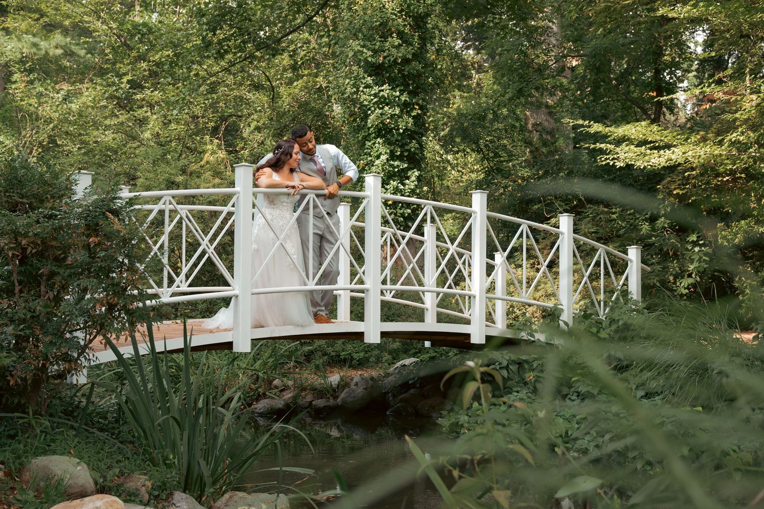 Couple sharing a romantic kiss on a white garden bridge surrounded by lush green trees.