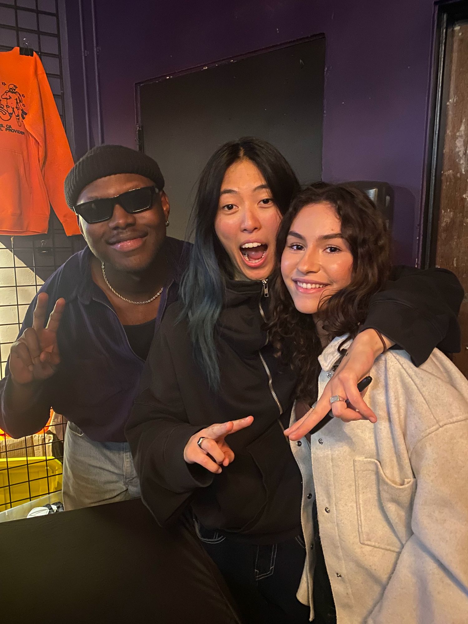 Three friends posing and smiling together indoors in a purple-walled venue.