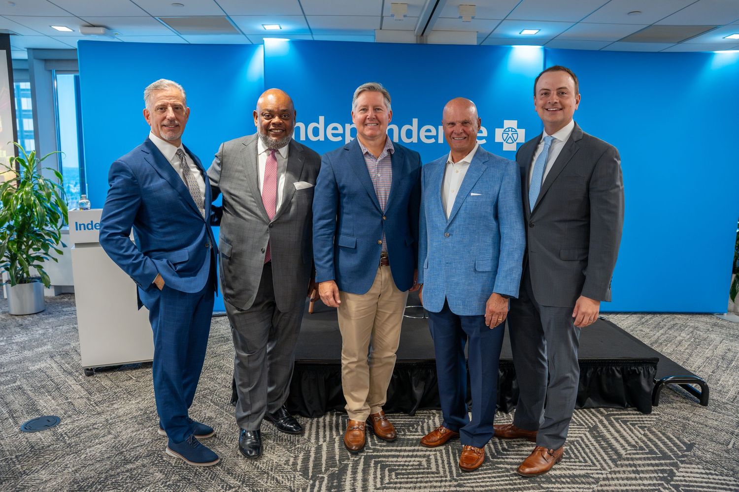 Five businessmen in suits pose together smiling in front of Independence Blue Cross branded backdrop.