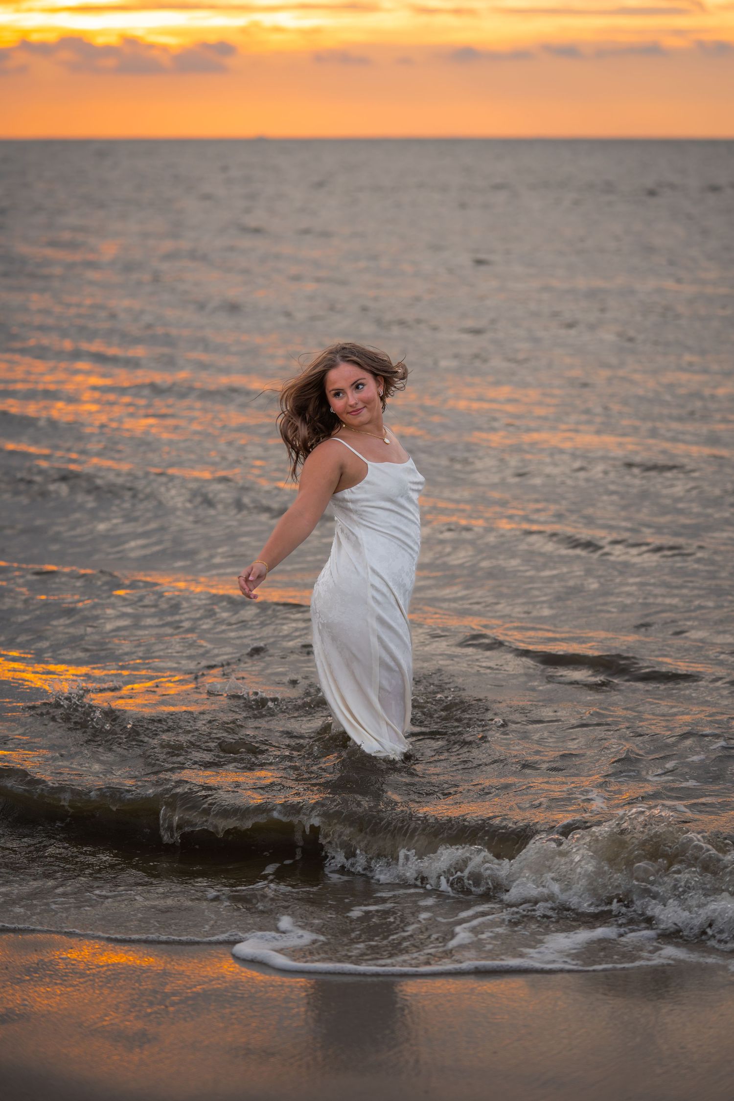 A young woman in a white dress wading in ocean waves during a golden sunset.