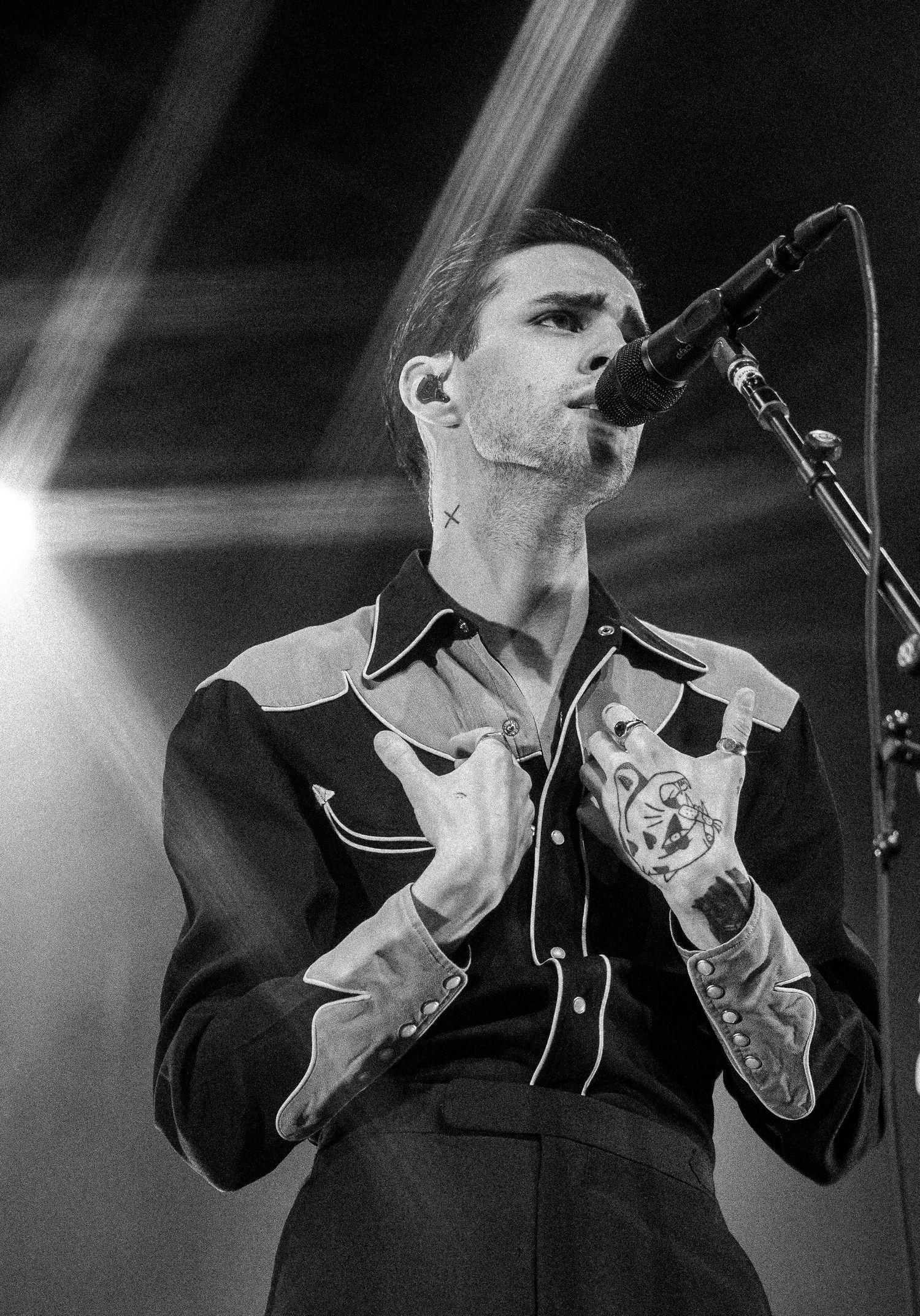 Black and white close-up of a tattooed male vocalist singing into a microphone under stage spotlights.