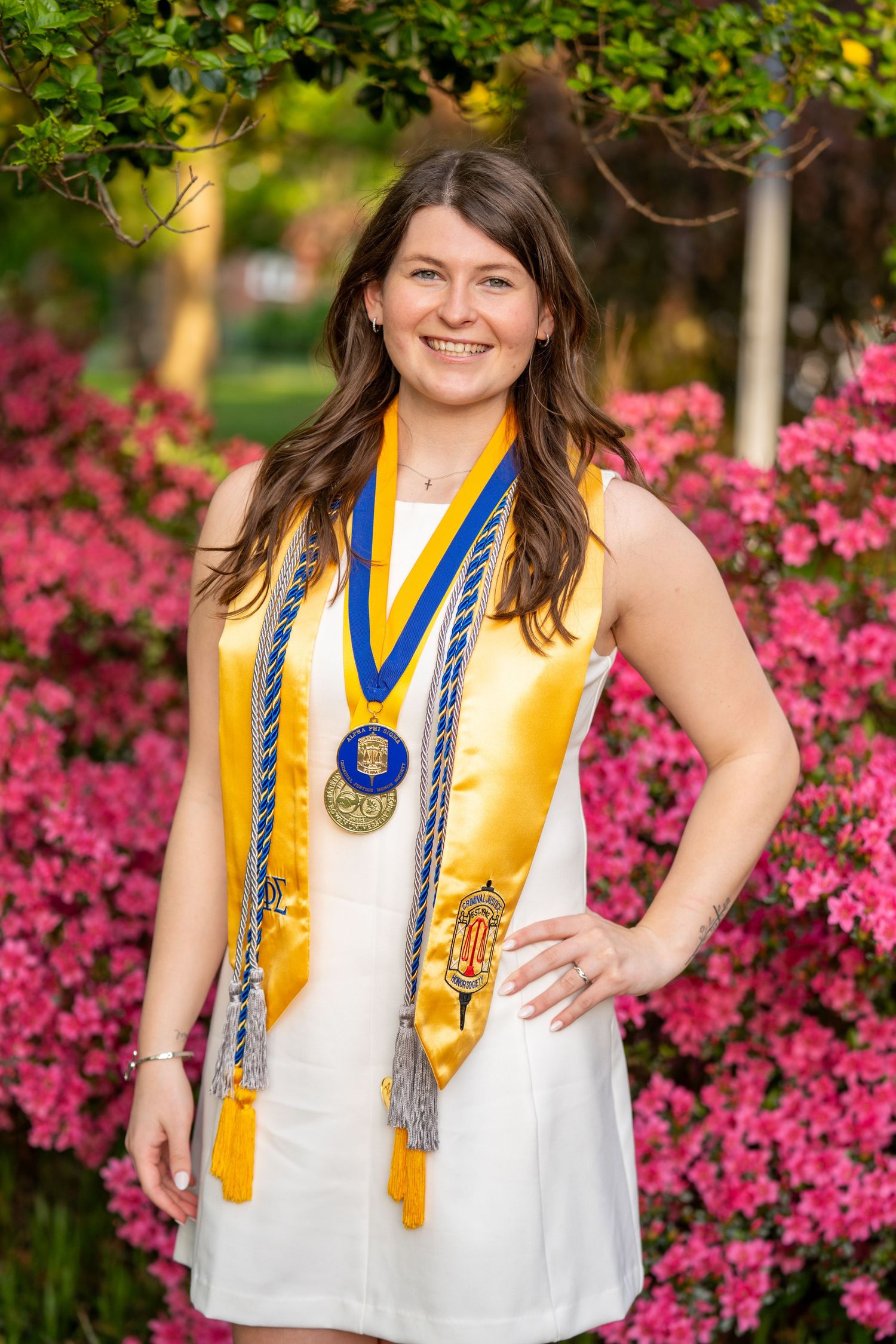 Smiling female graduate wearing gold sash, medals, and blue honor cords poses in front of pink flowering bushes.
