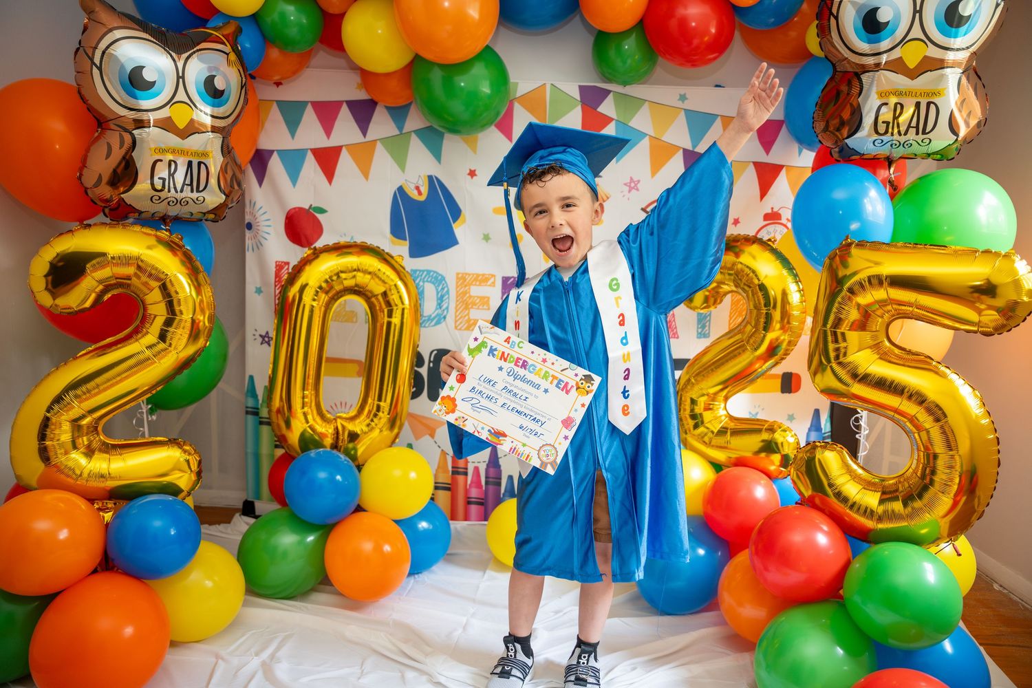 Happy child in blue graduation cap and gown holding diploma at colorful 2025 graduation party.