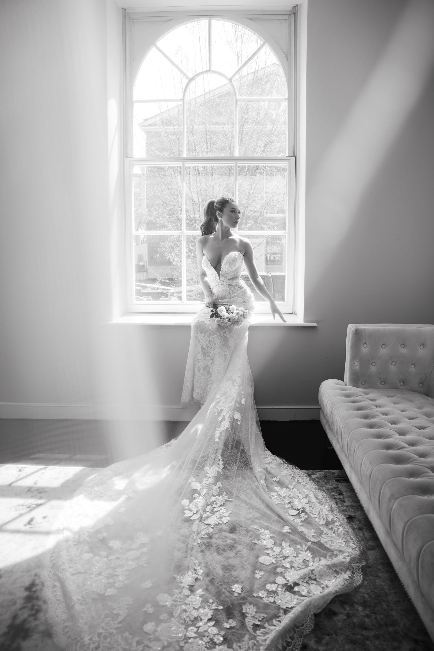 Black and white full-length portrait of bride in lace gown with train standing by arched window.