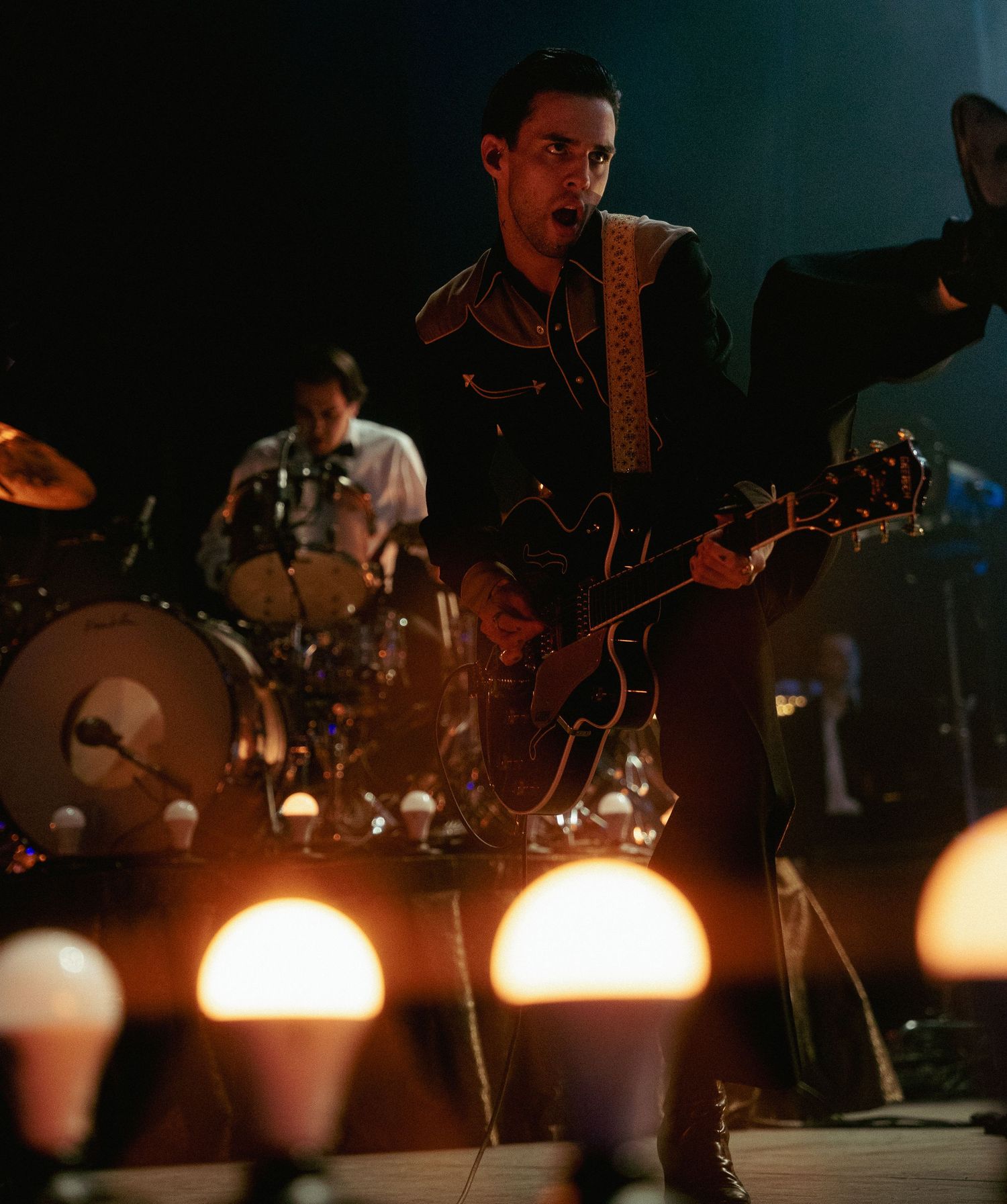Male guitarist playing a dark semi-hollow guitar on a dimly lit stage with glowing lights in foreground.