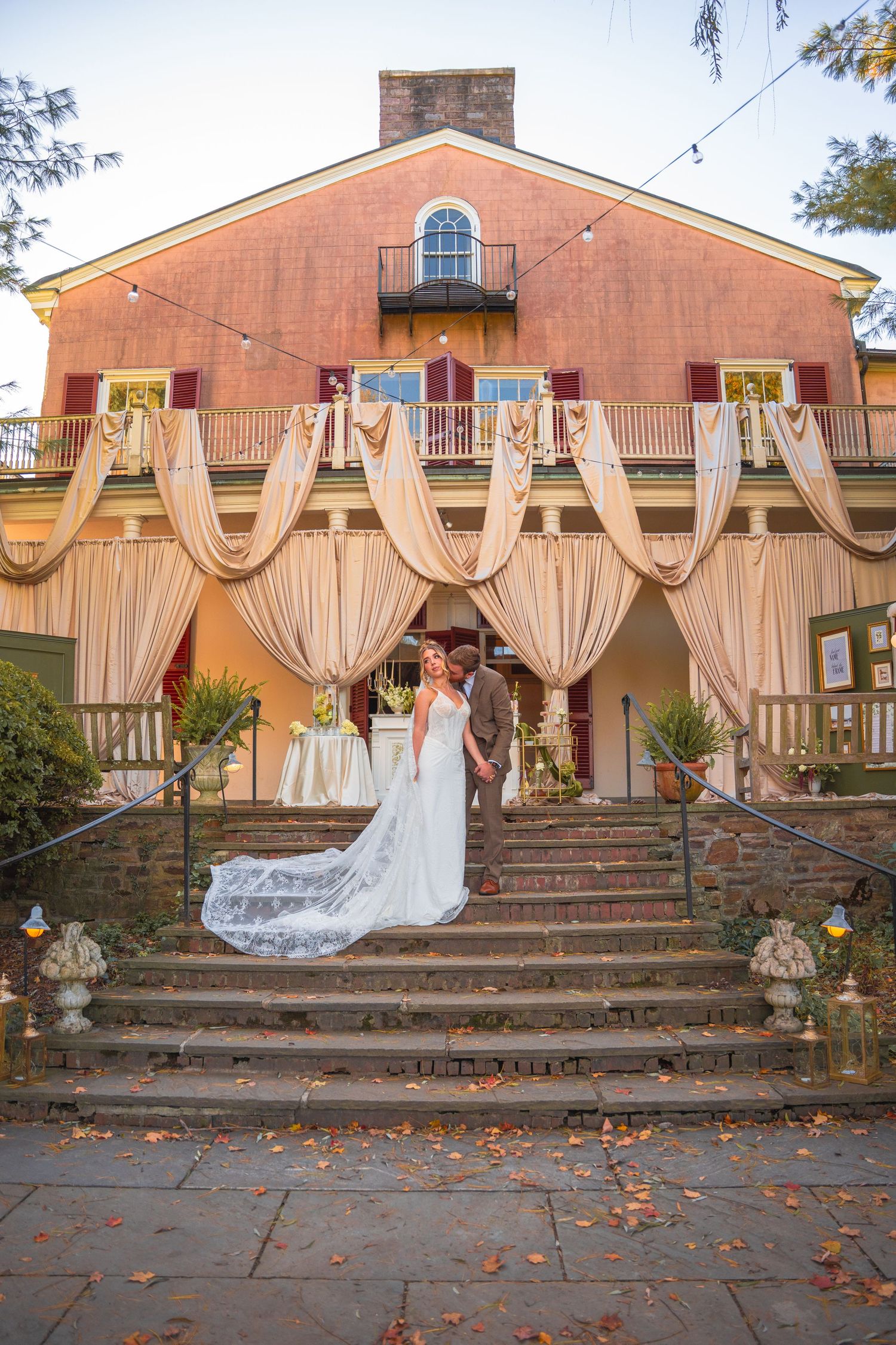Bride and groom pose on steps of a decorated historic venue with draped fabric at sunset.