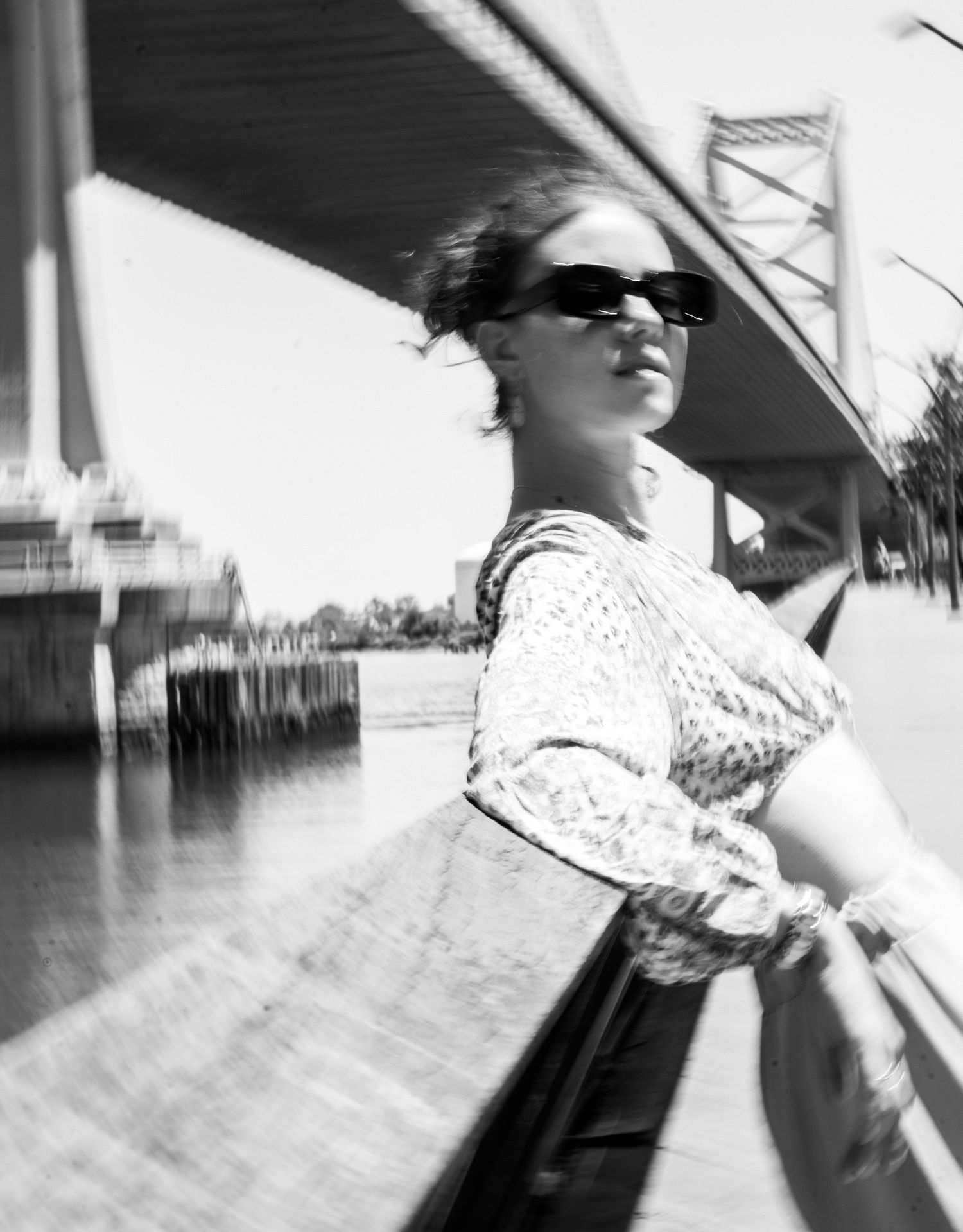 Black and white portrait of woman in sunglasses leaning on railing with bridge in background.