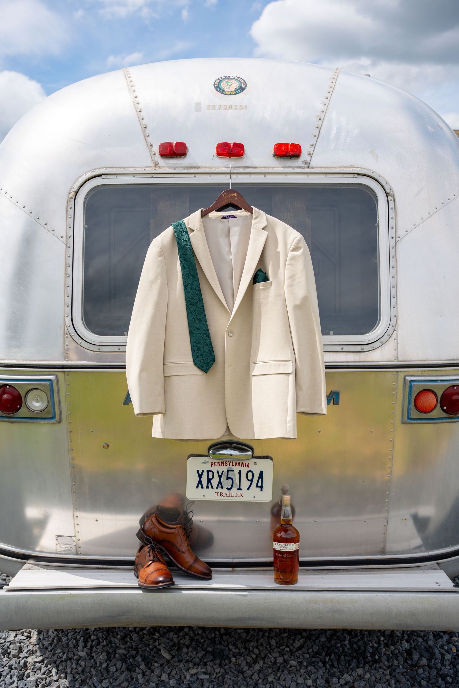White wedding suit with green tie on hanger displayed on vintage Airstream trailer with brown dress shoes and whiskey bottle.