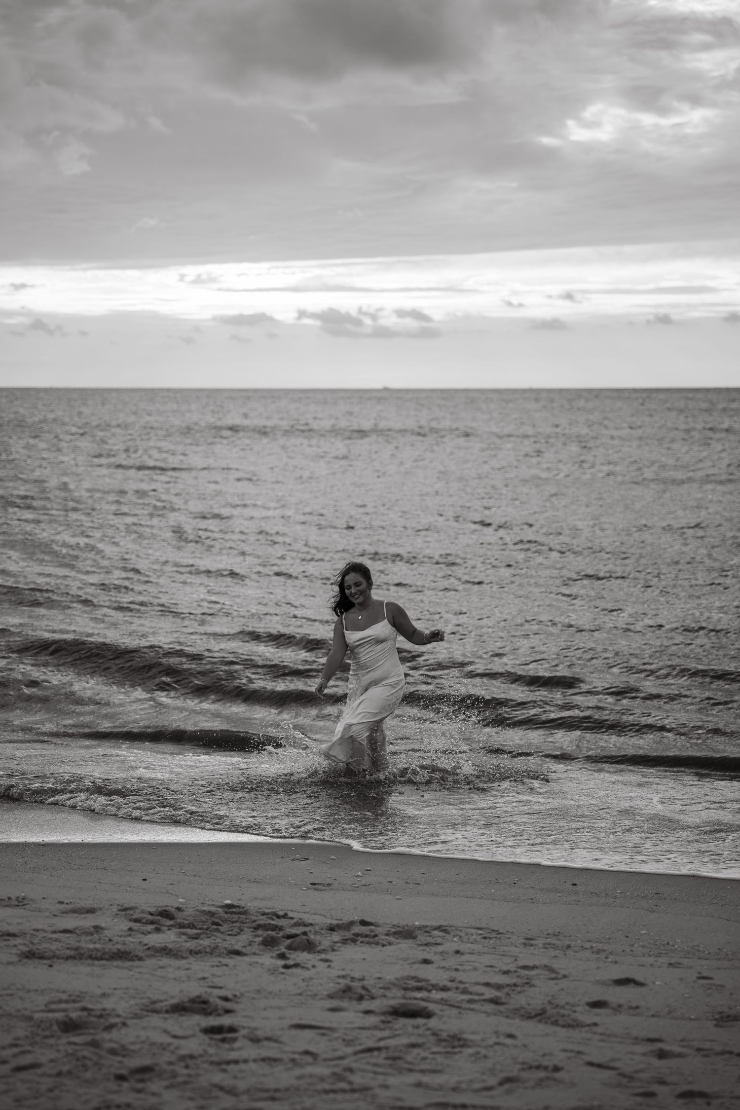 Black and white photo of woman in white dress running along ocean shoreline at dusk.