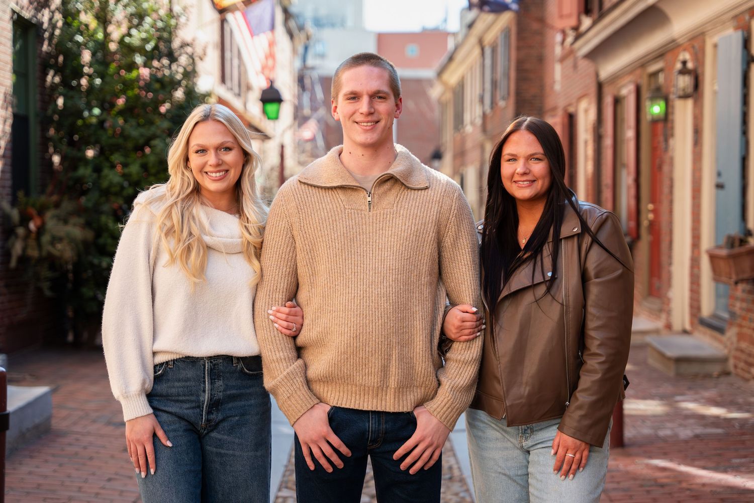 Three smiling young adults posing together on a charming brick-lined street outdoors.