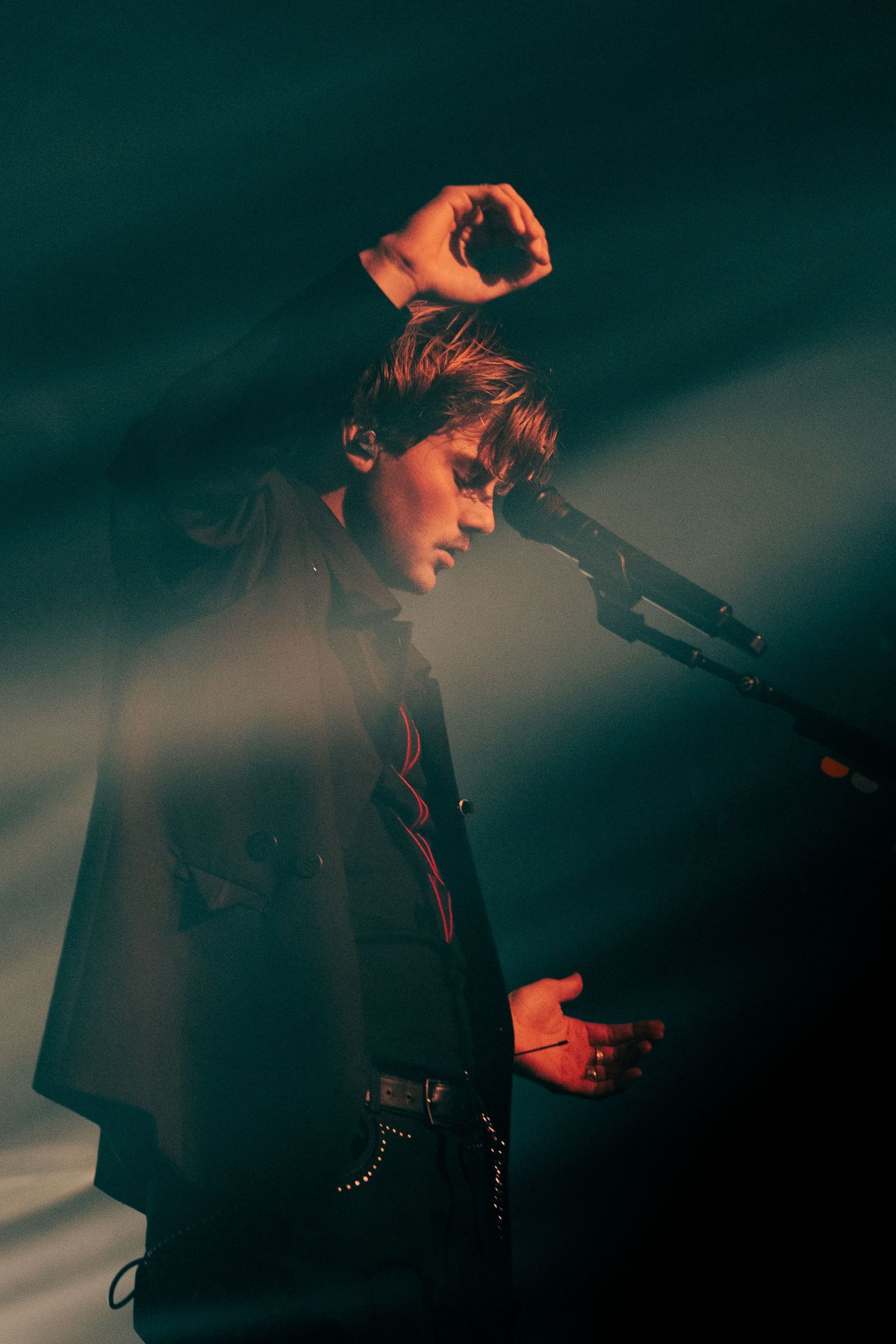 Male performer on stage in dramatic cape coat under teal and red spotlights at microphone.