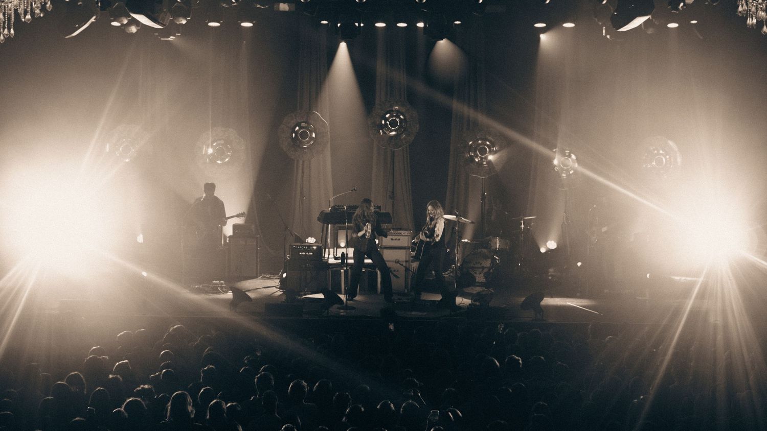 Wide-angle sepia-toned shot of a full band performing on a warmly lit stage before a packed audience.