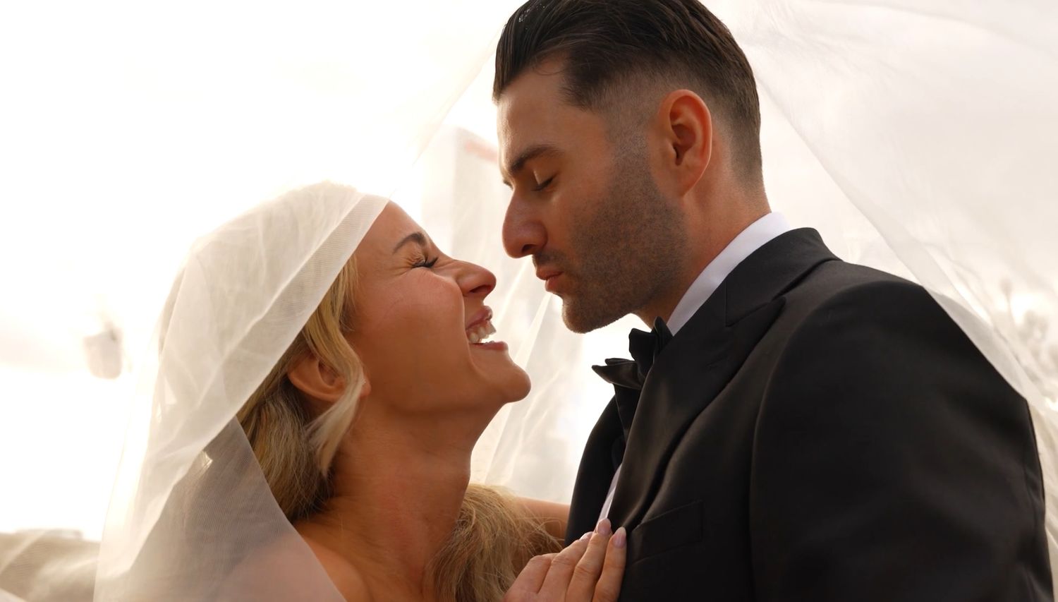 Bride and groom sharing a tender moment under a flowing white veil in soft romantic lighting.