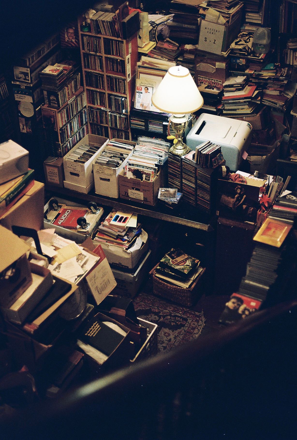 A cluttered room filled with stacks of books, records, and boxes illuminated by a lamp.