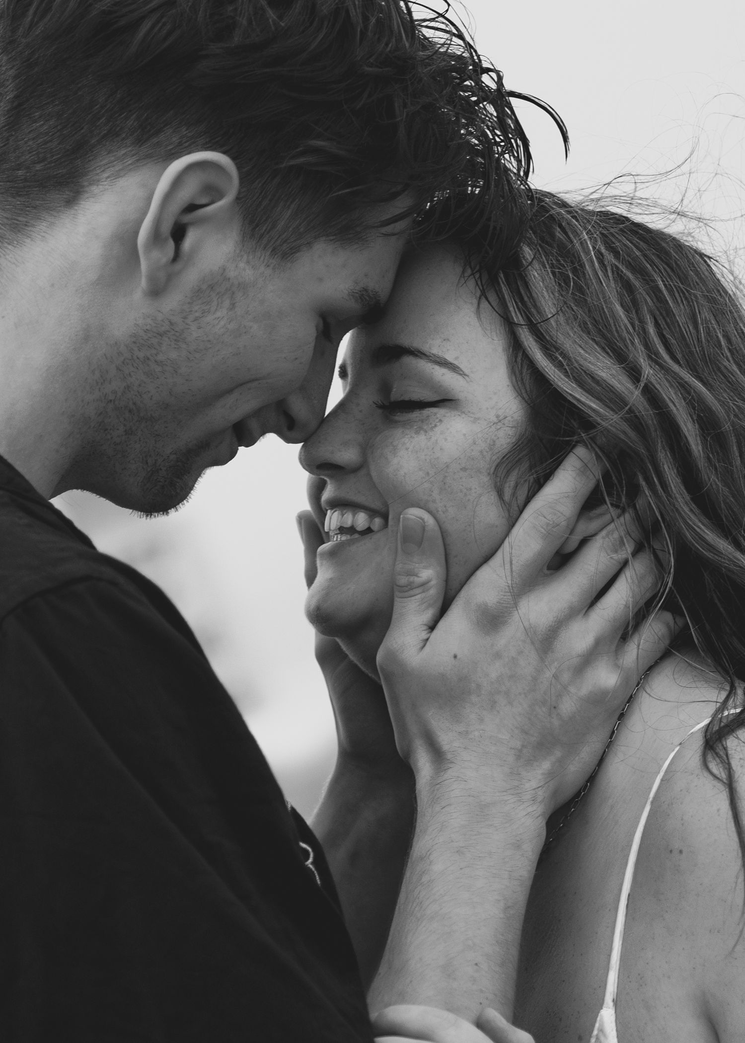 Black and white close-up of a smiling couple touching foreheads tenderly on a beach.