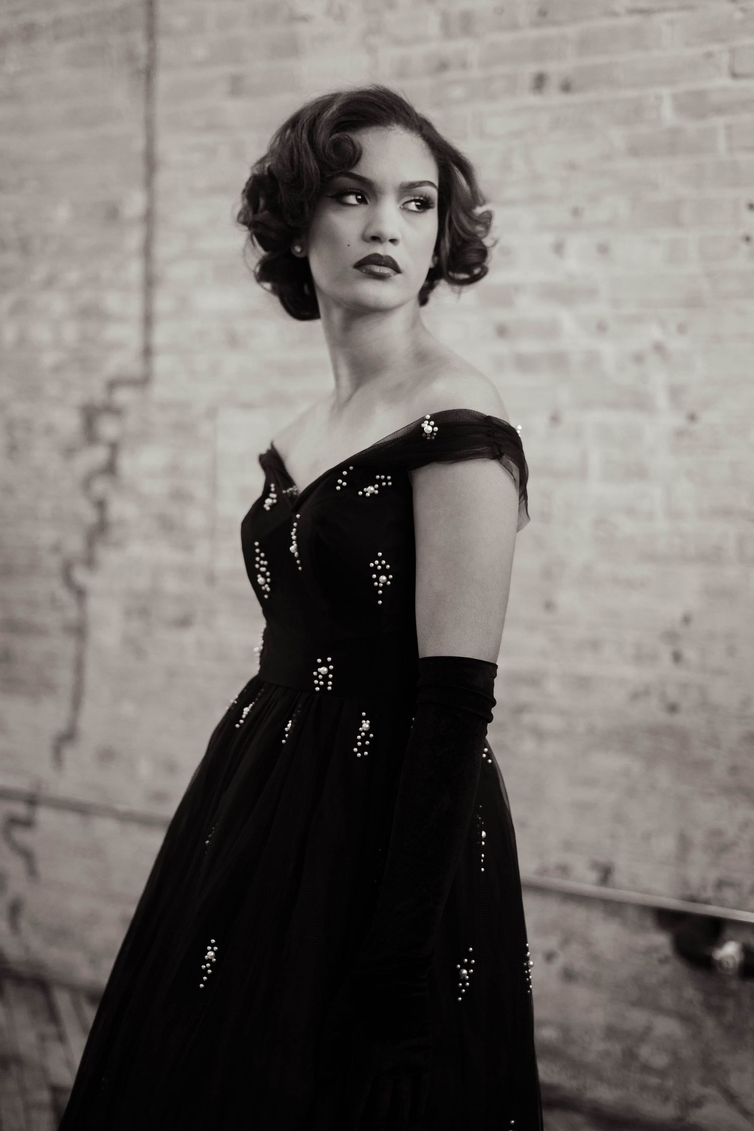 Elegant woman in black sequin off-shoulder gown posing before brick wall in black and white.