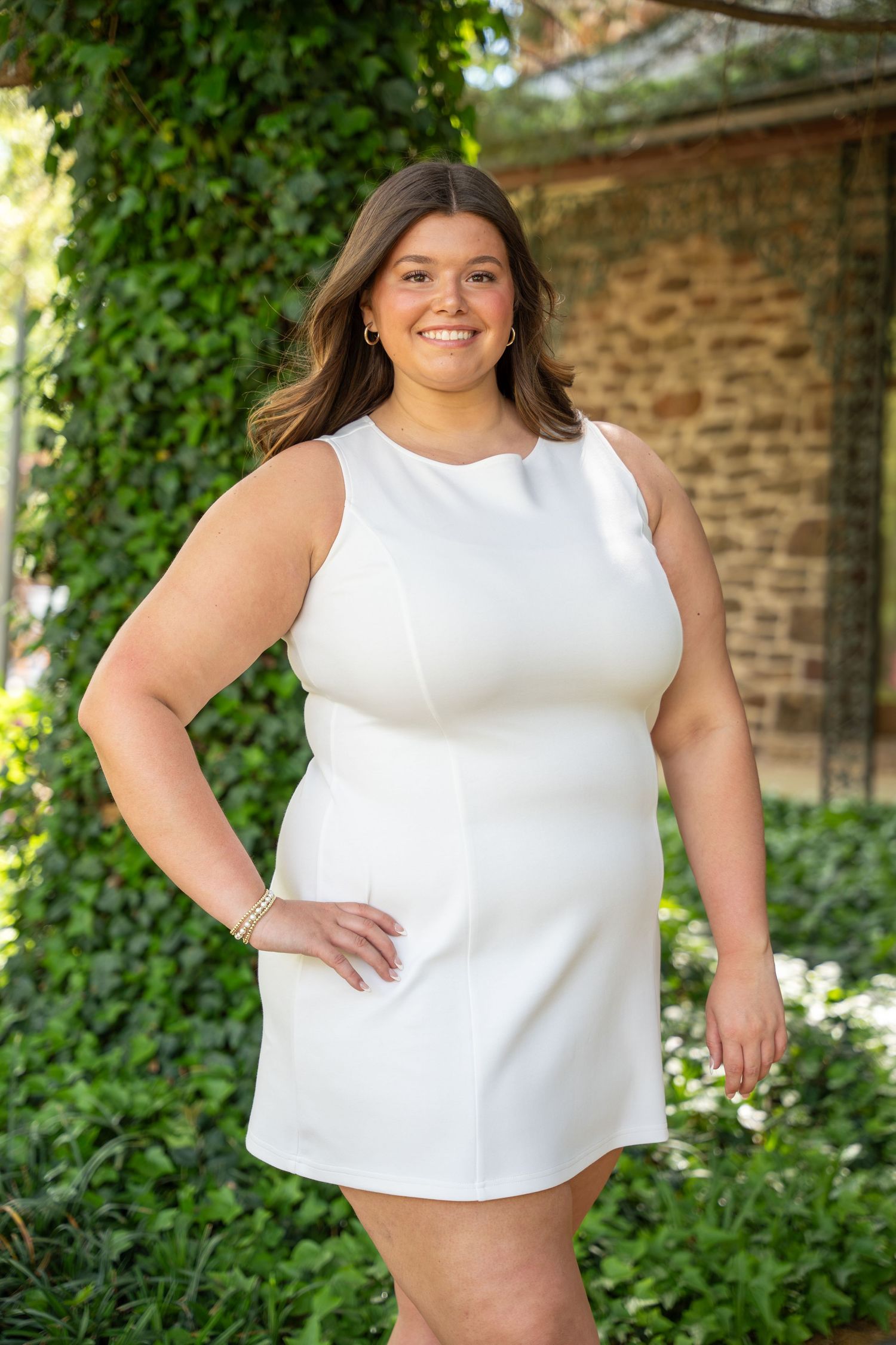 Smiling young woman in white sleeveless dress posing outdoors against ivy-covered stone wall.
