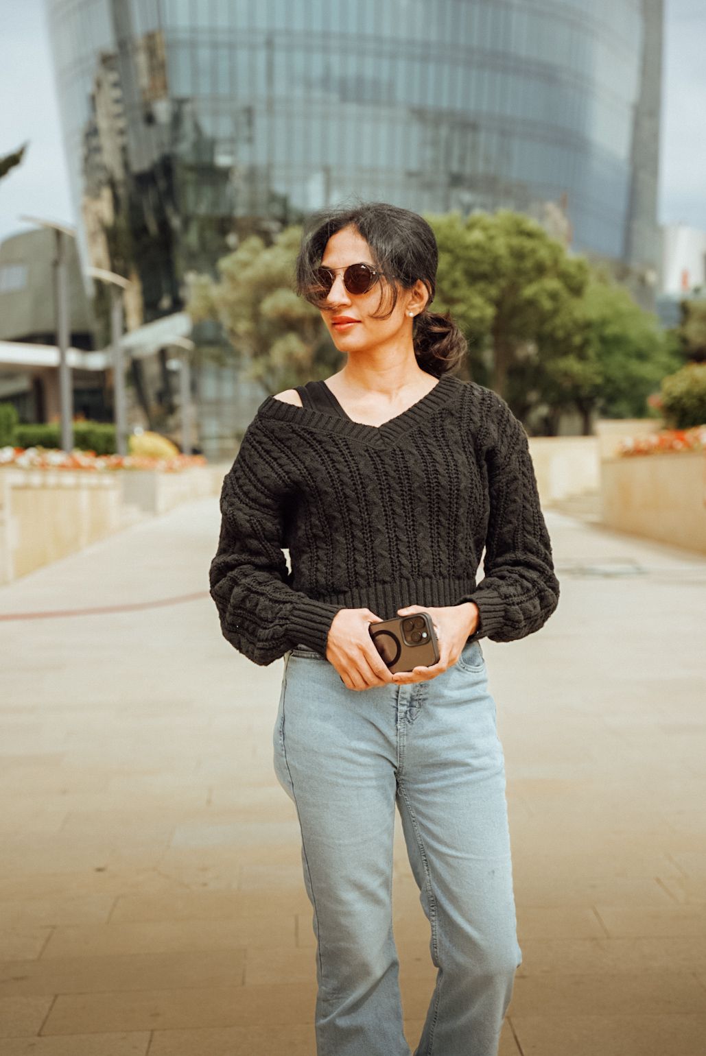 Person wearing a black cable knit sweater and light jeans poses in an urban setting.