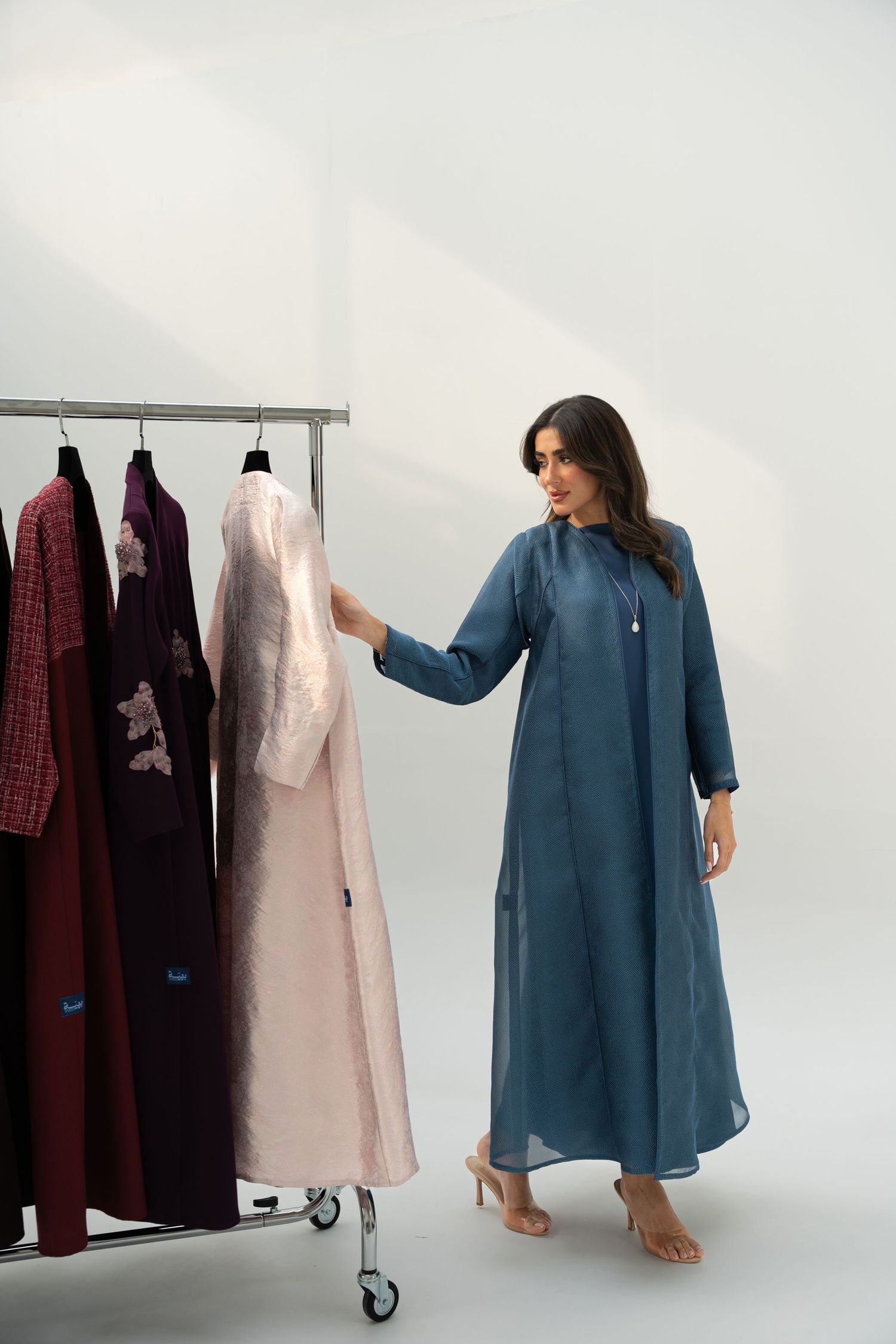 Model wearing a teal blue tunic dress poses with clothing rack displaying burgundy and pink garments in studio setting.