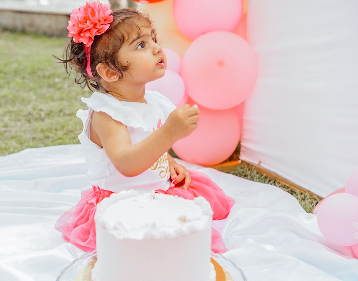 Baby girl in white dress plays with pink balloons at outdoor celebration.