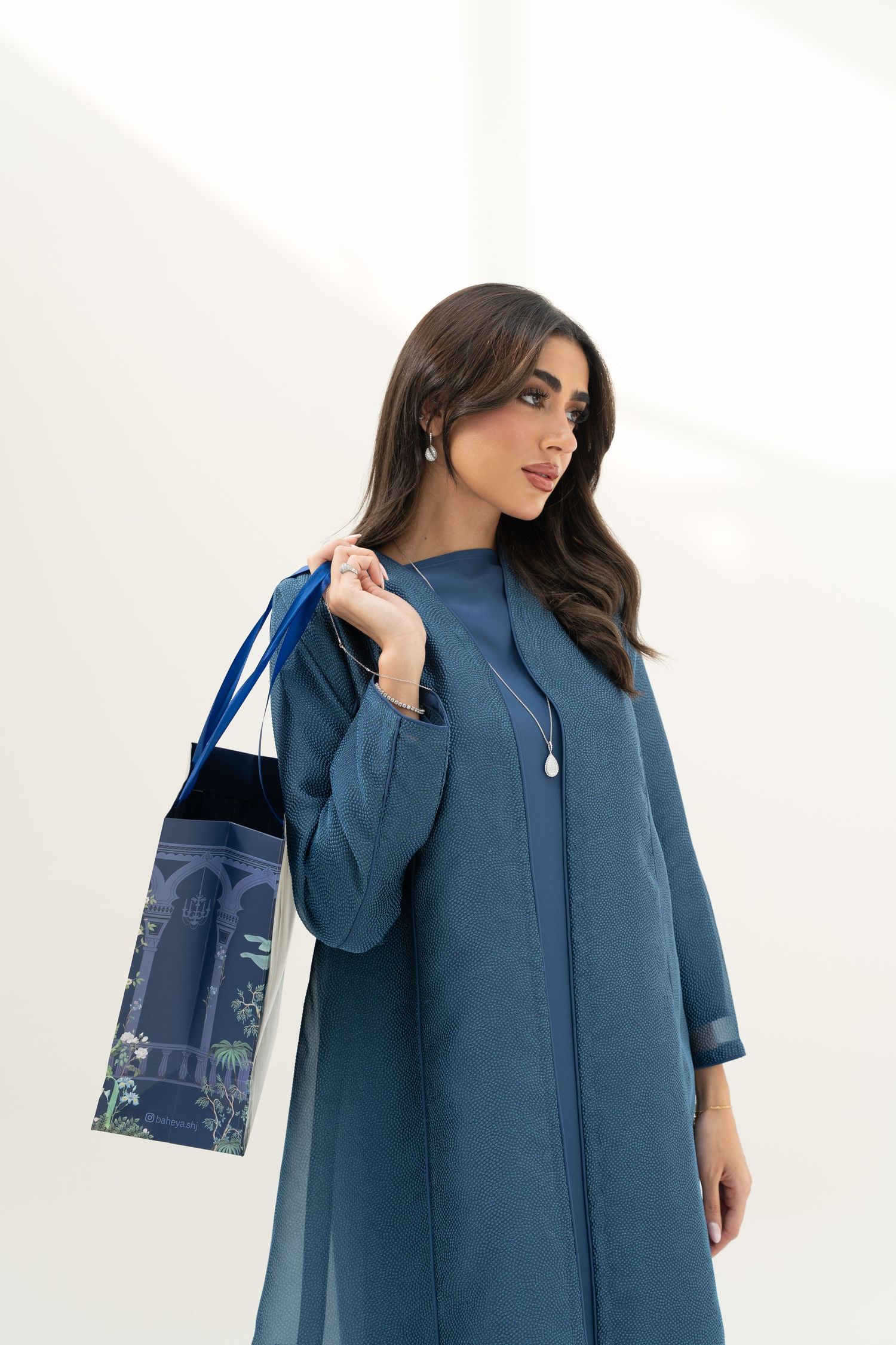 Woman in elegant teal blue abaya dress poses with matching navy tote bag against white wall.
