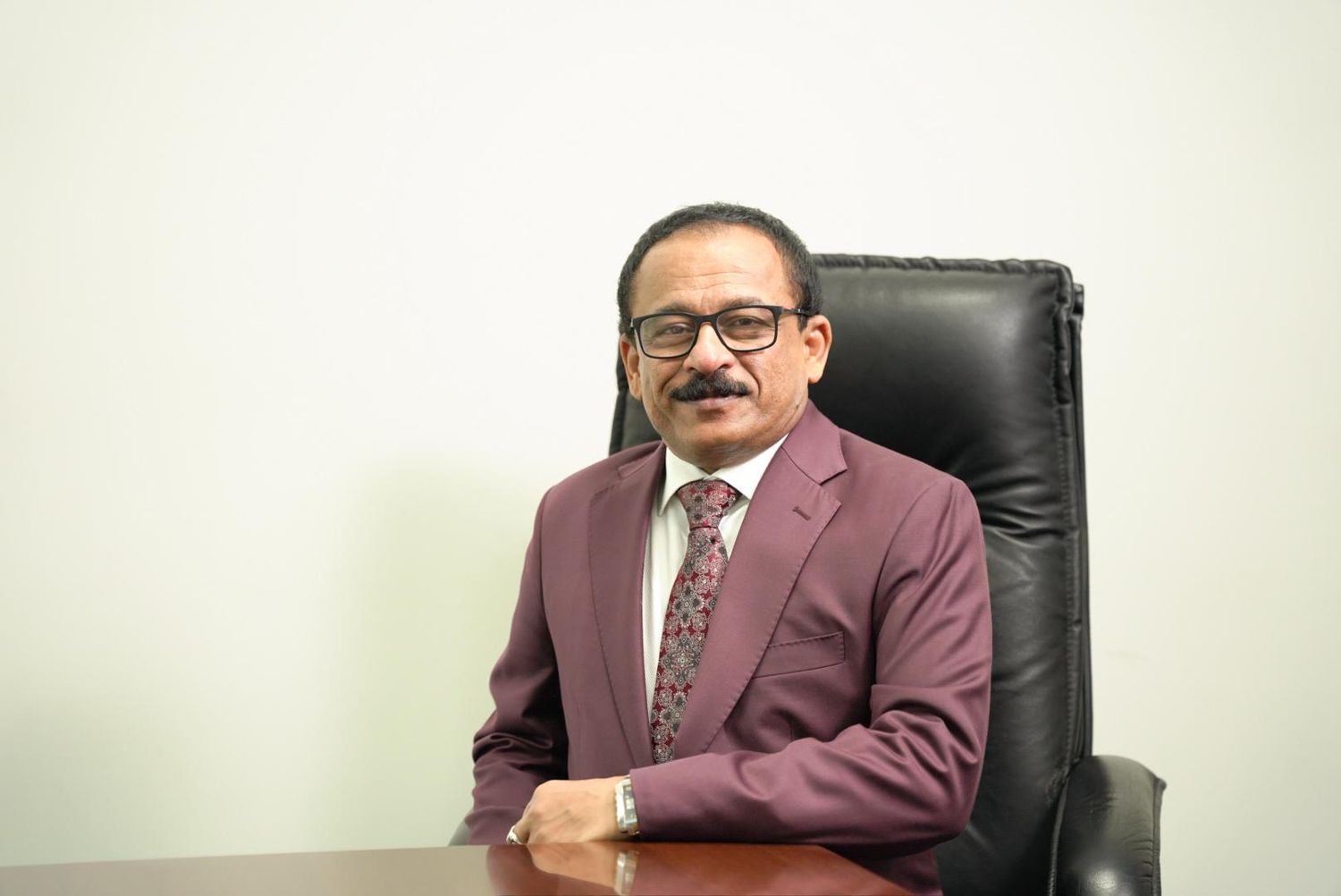 A professional in a burgundy suit jacket and patterned tie sits at a desk in an office setting.