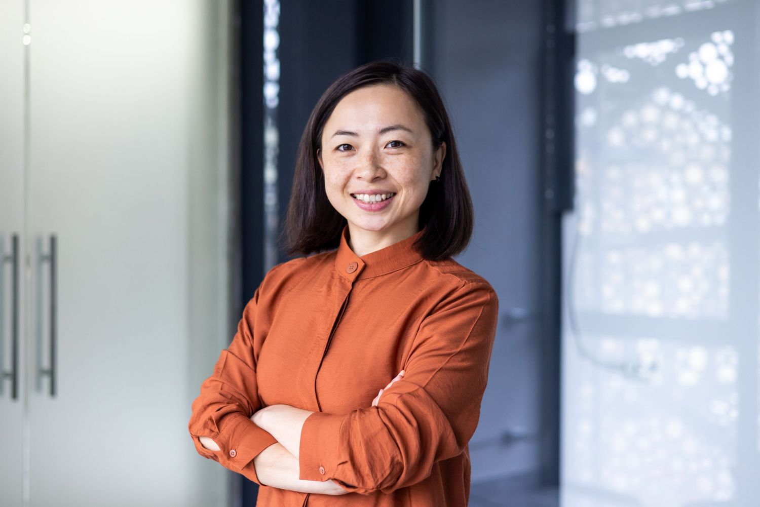 Woman in orange blazer poses confidently in modern office setting.