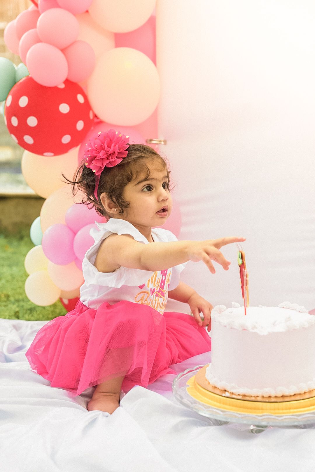 Baby in pink tutu dress reaches for birthday cake at festive celebration.