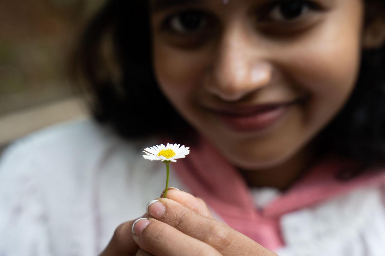 A child holds a small white daisy flower while smiling outdoors in natural lighting.