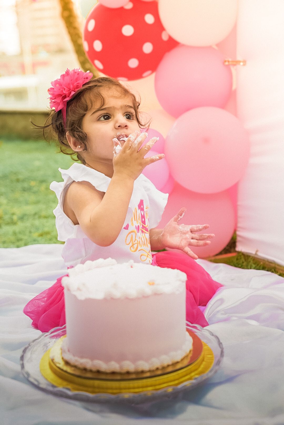 Birthday celebration with pink balloons and white frosted cake on picnic blanket.