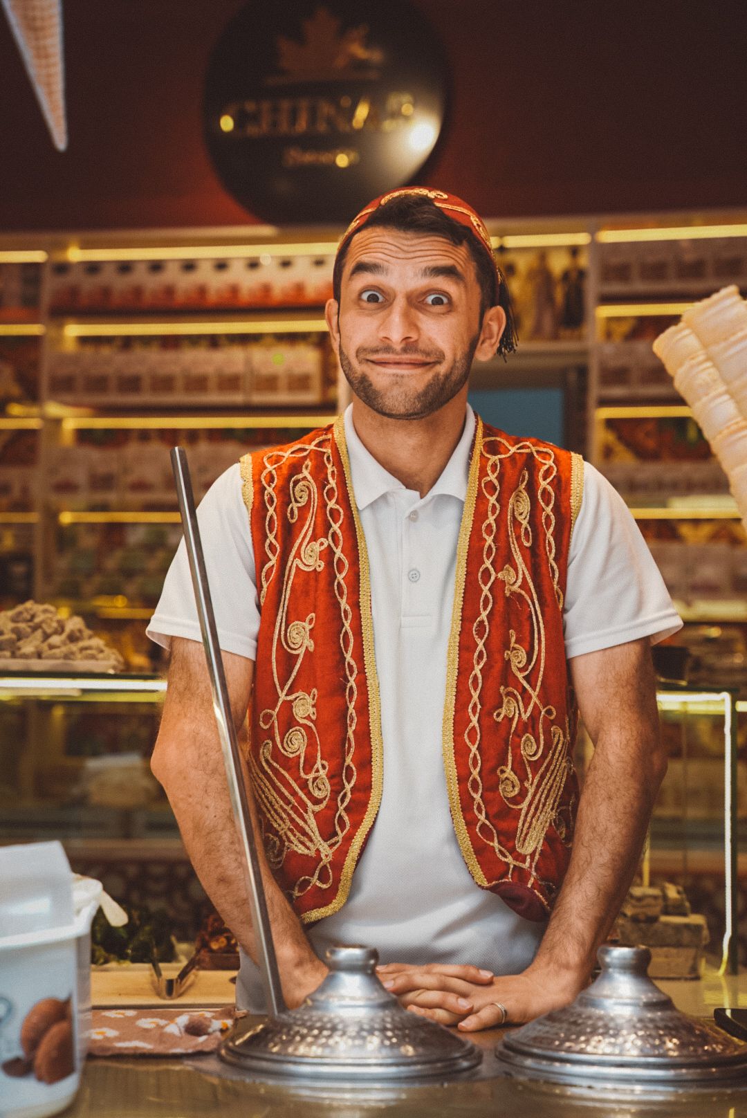 Ice cream vendor in traditional vest smiles behind glass display counter.