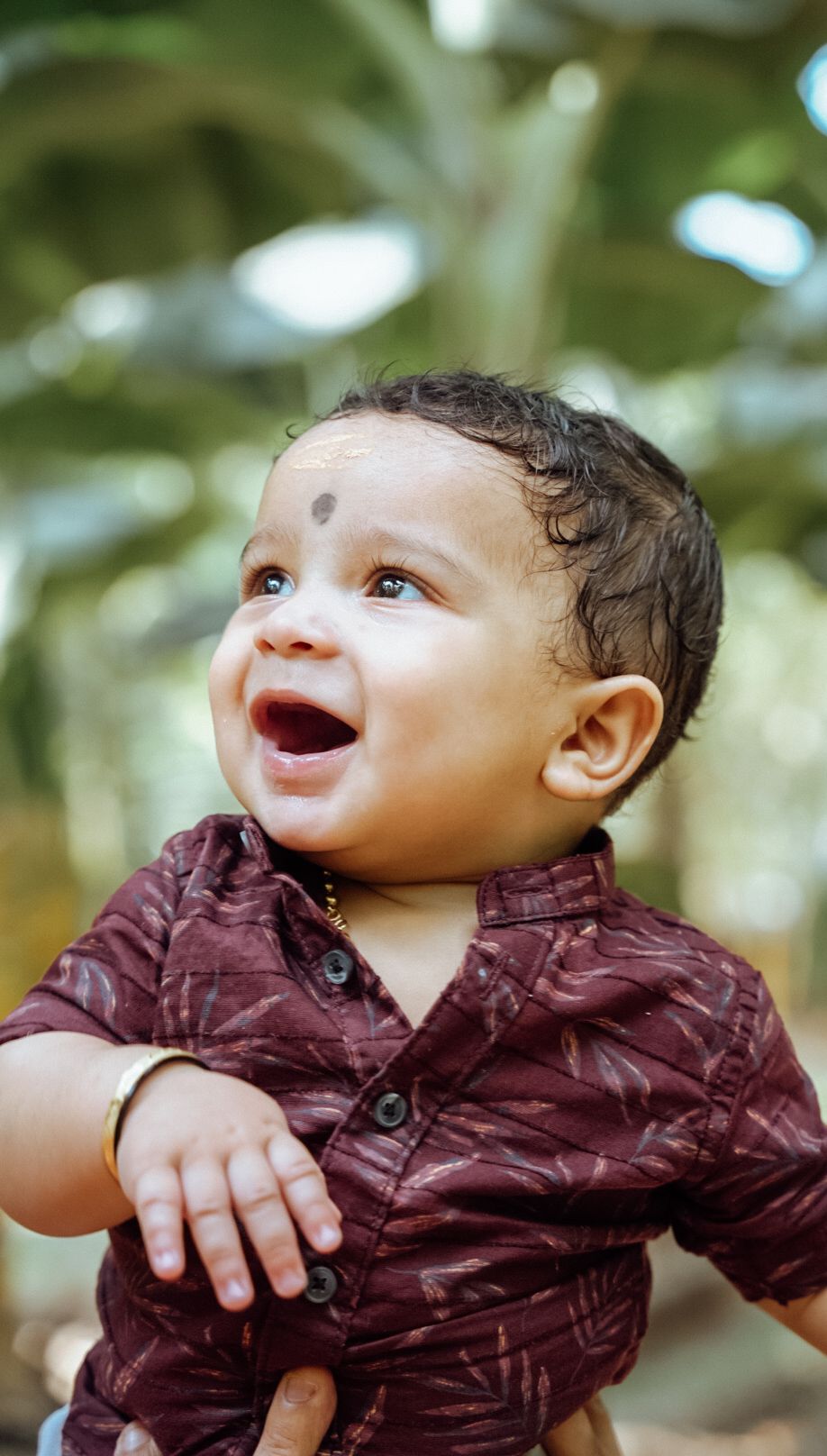 Baby in burgundy shirt showing expressions of wonder and joy while looking up at nature.