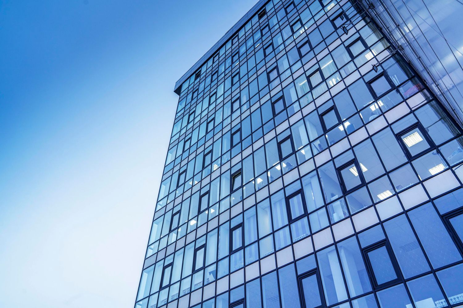 Modern glass office building facade photographed from below against blue sky.