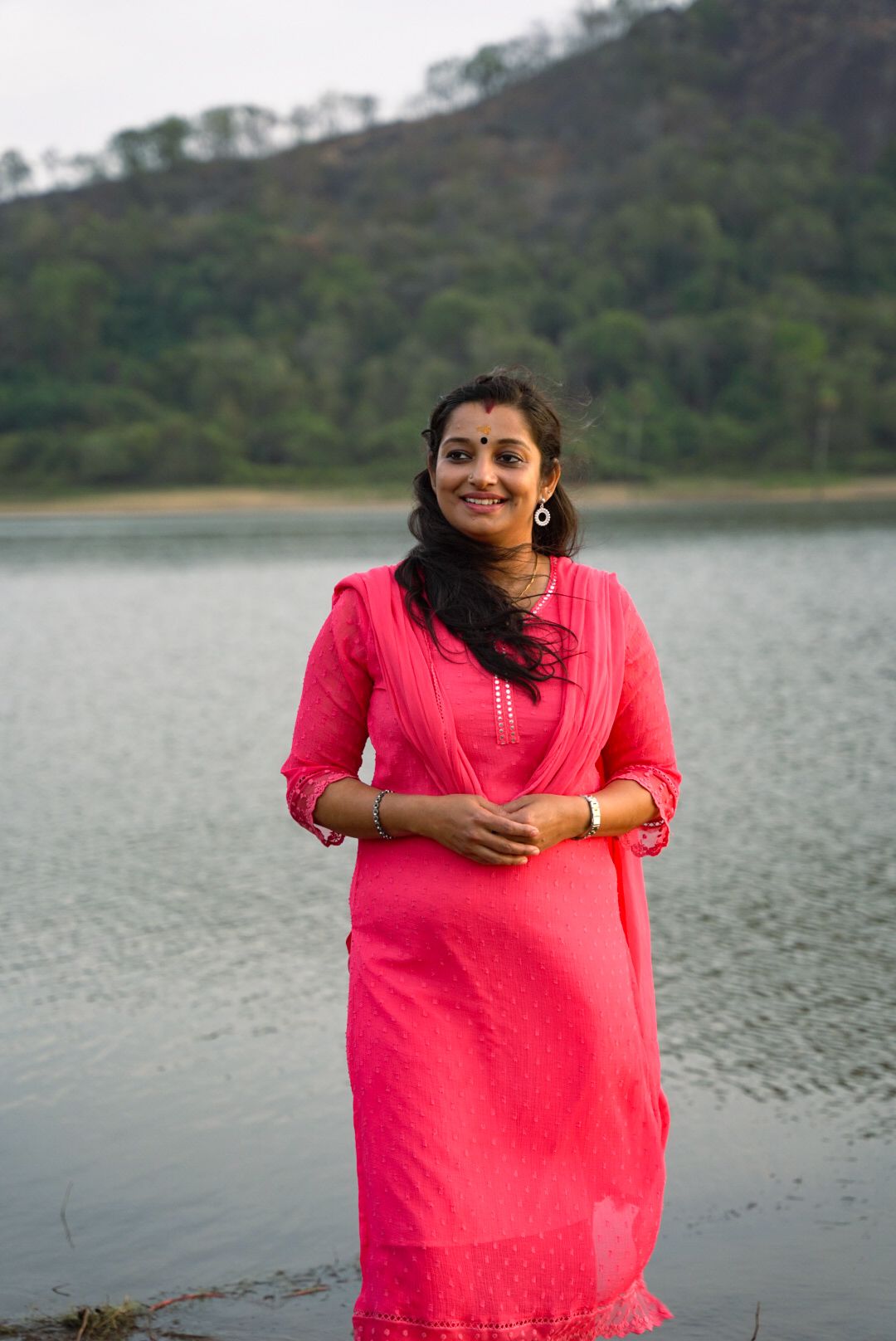 Person in bright pink attire posing by a serene lake with mountains in the background.