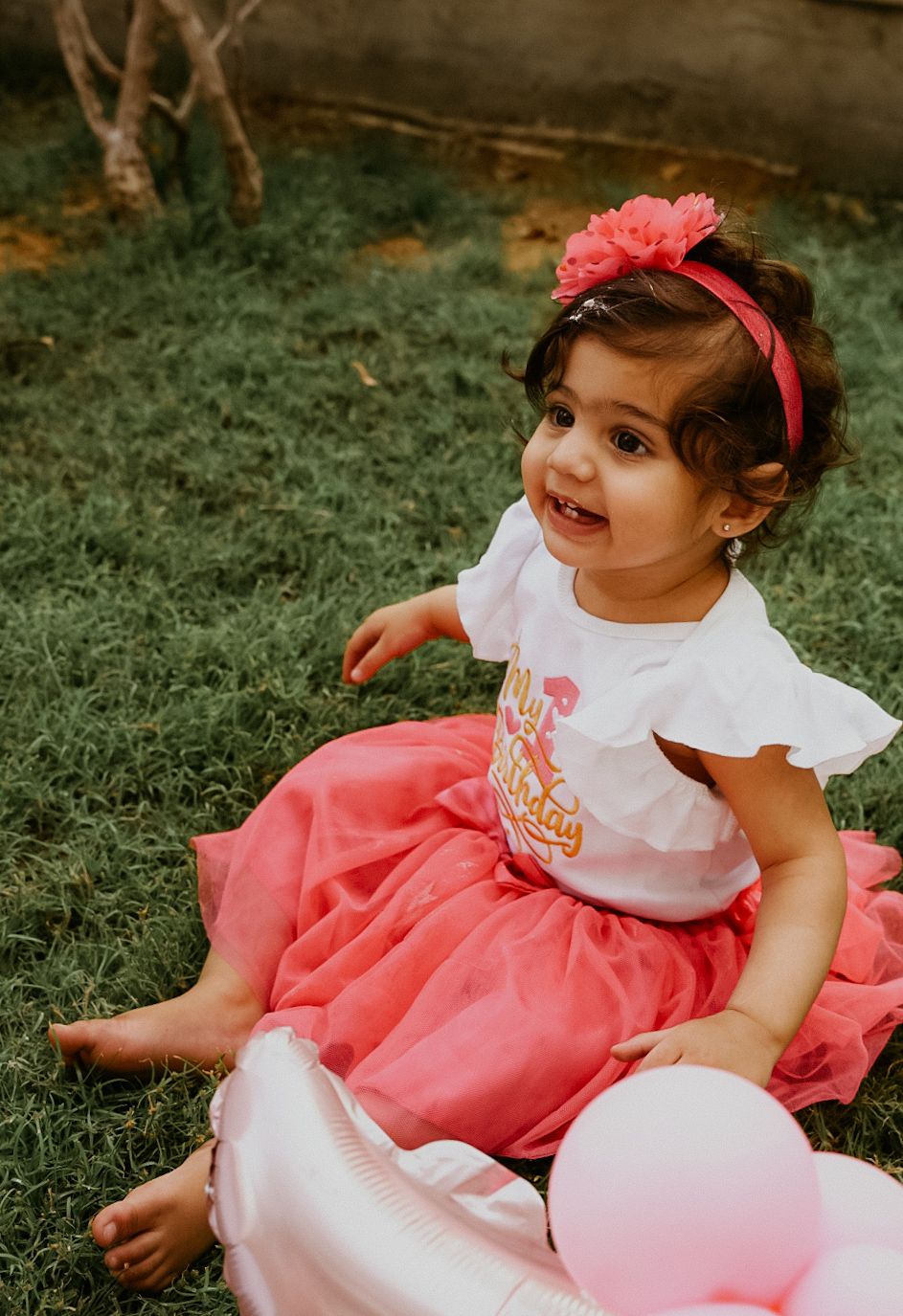 Baby wearing a pink tulle dress and white top sits on grass during a photo session.