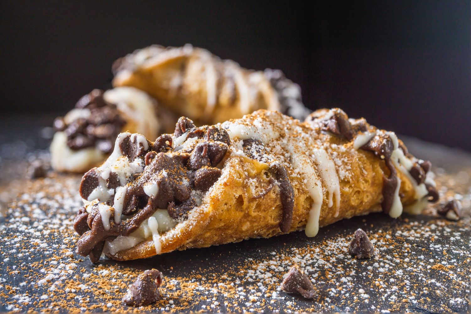 Freshly baked pastries with chocolate chips and powdered sugar on dark surface.