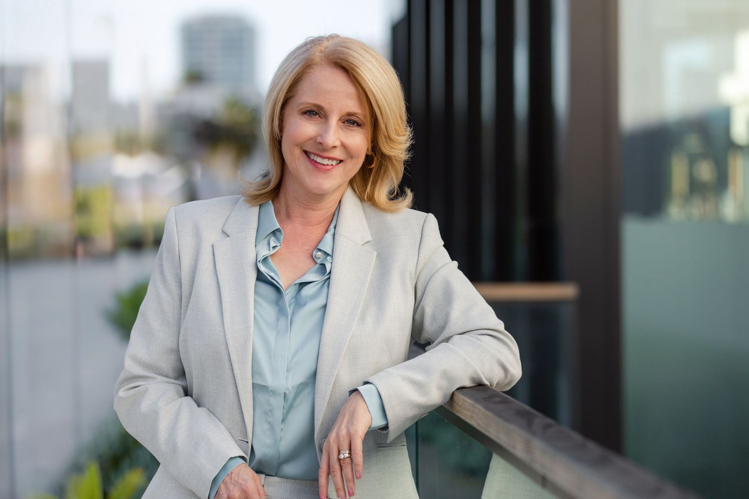 Professional in a light gray blazer poses outdoors with modern architecture in background.