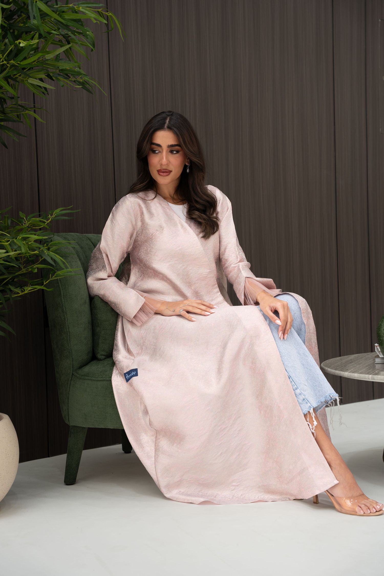 Elegant model in pale pink satin dress and light denim sits gracefully on dark green velvet accent chair.