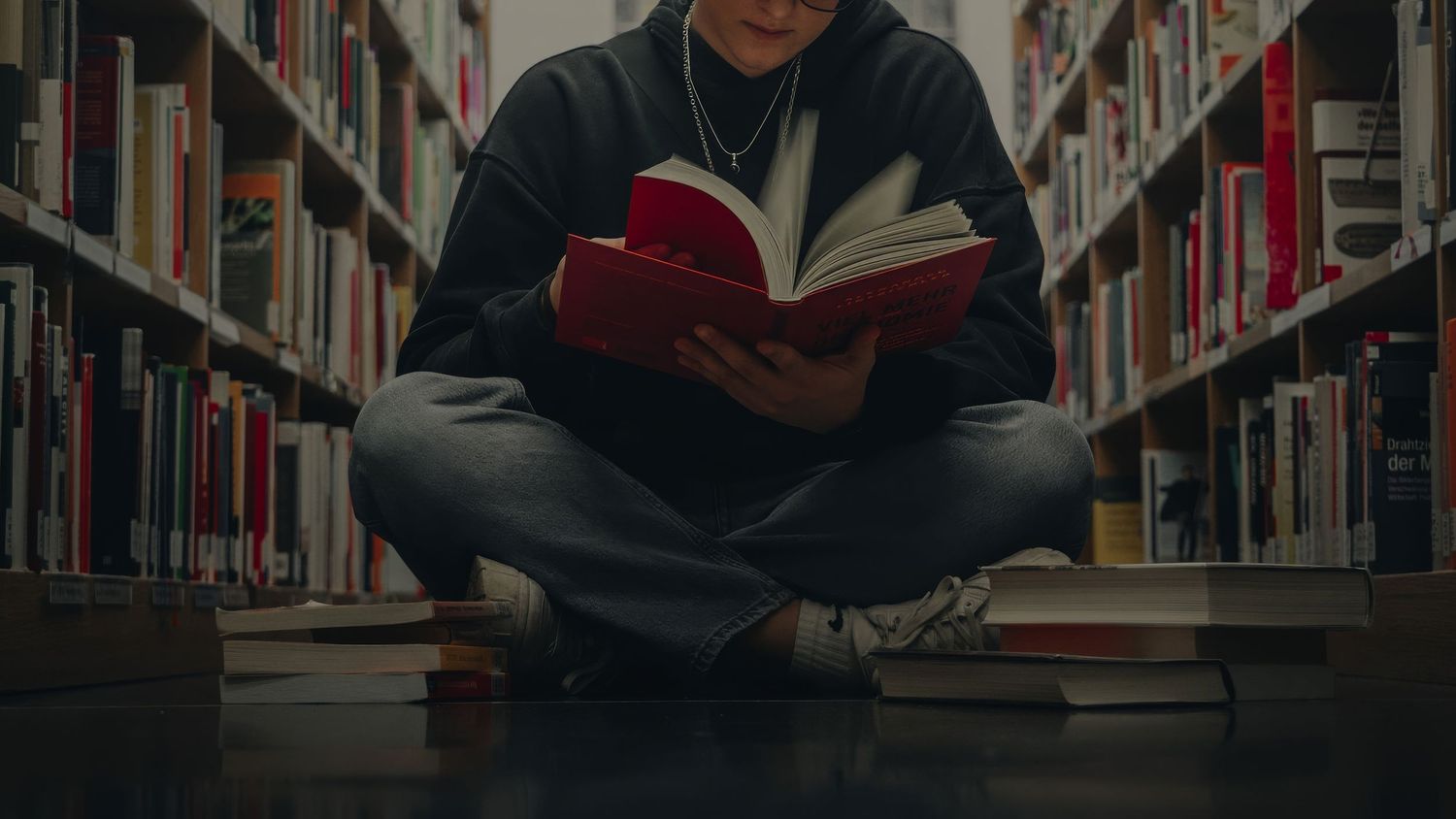 Someone sitting cross-legged reading a red book between library shelves in moody lighting.